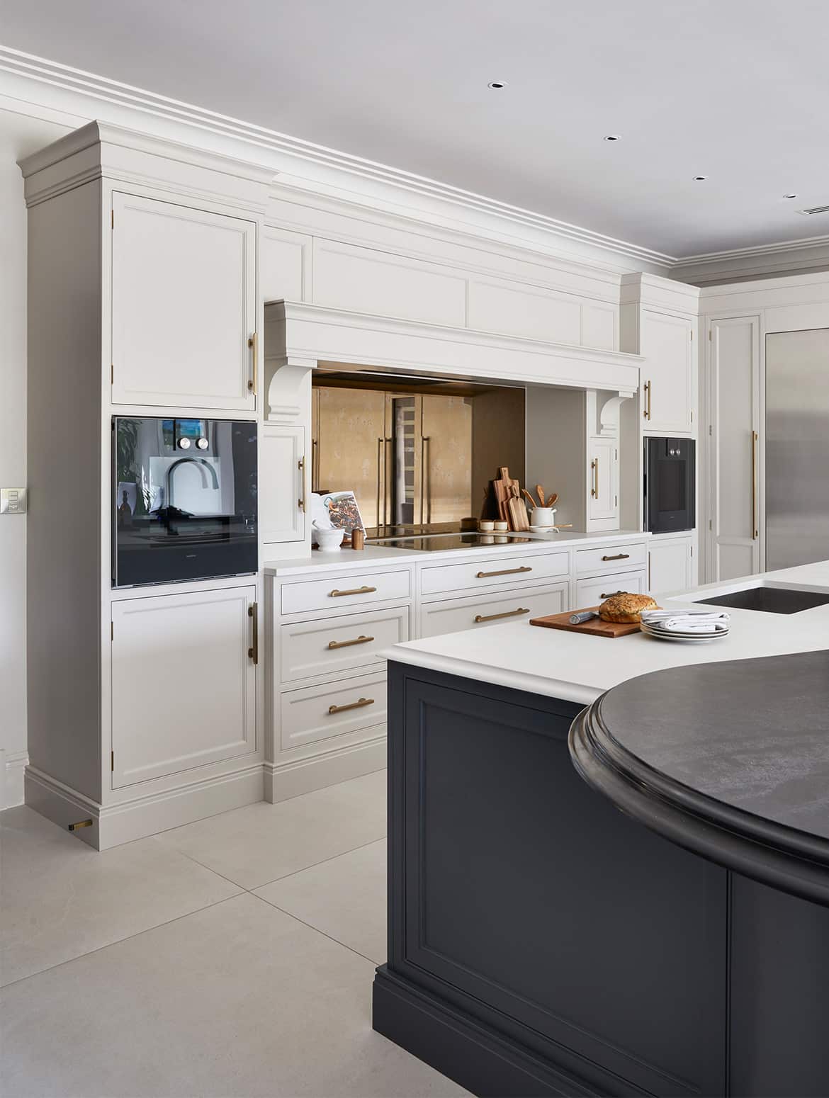 A traditional shaker kitchen project in off-black. A central island with the bespoke cabinetry and a streamlined wood ledge contrasts the white worktop of the remaining island beyond. To its right, two Gaggenau ovens are built-in to the middle French Grey cabinetry. The mantel stands proud to the surrounding cabinetry. 