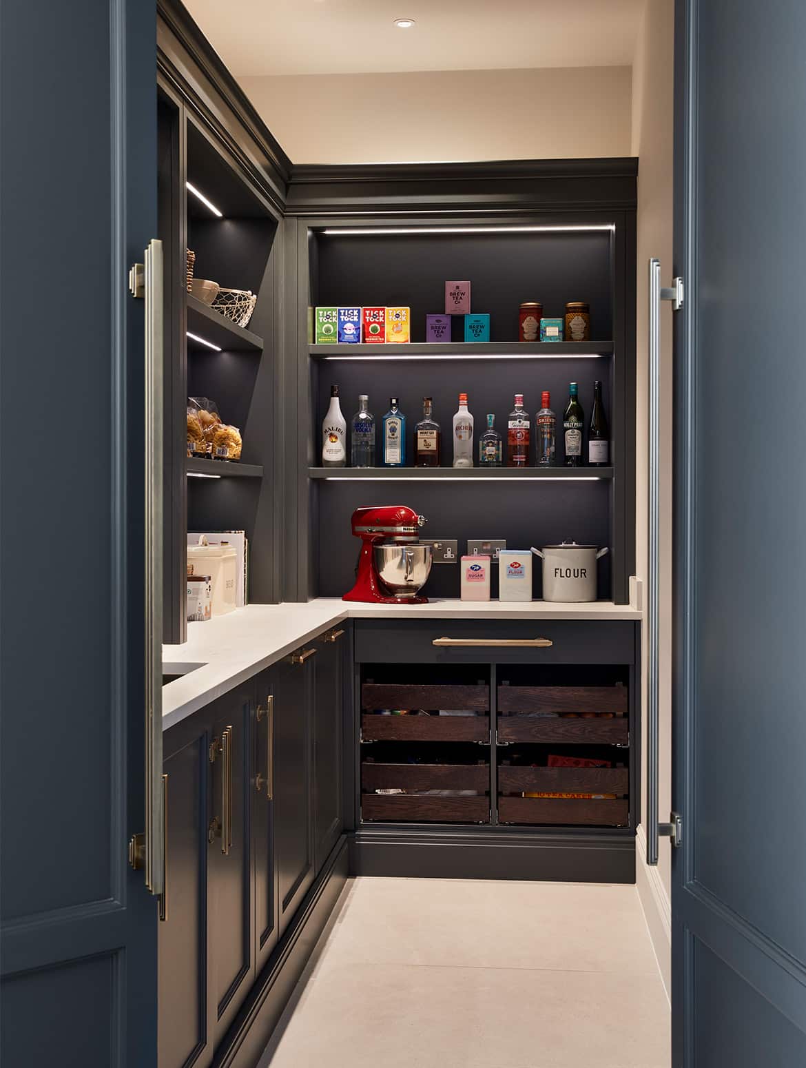 A traditional shaker kitchen project in off-black. The open doors of a hidden pantry that has bespoke off-black cabinetry above and below a white worktop. The worktop has a baking station with a red stand mixer and flour next to it. On the other side, there is a store of savoury goods such as pasta.