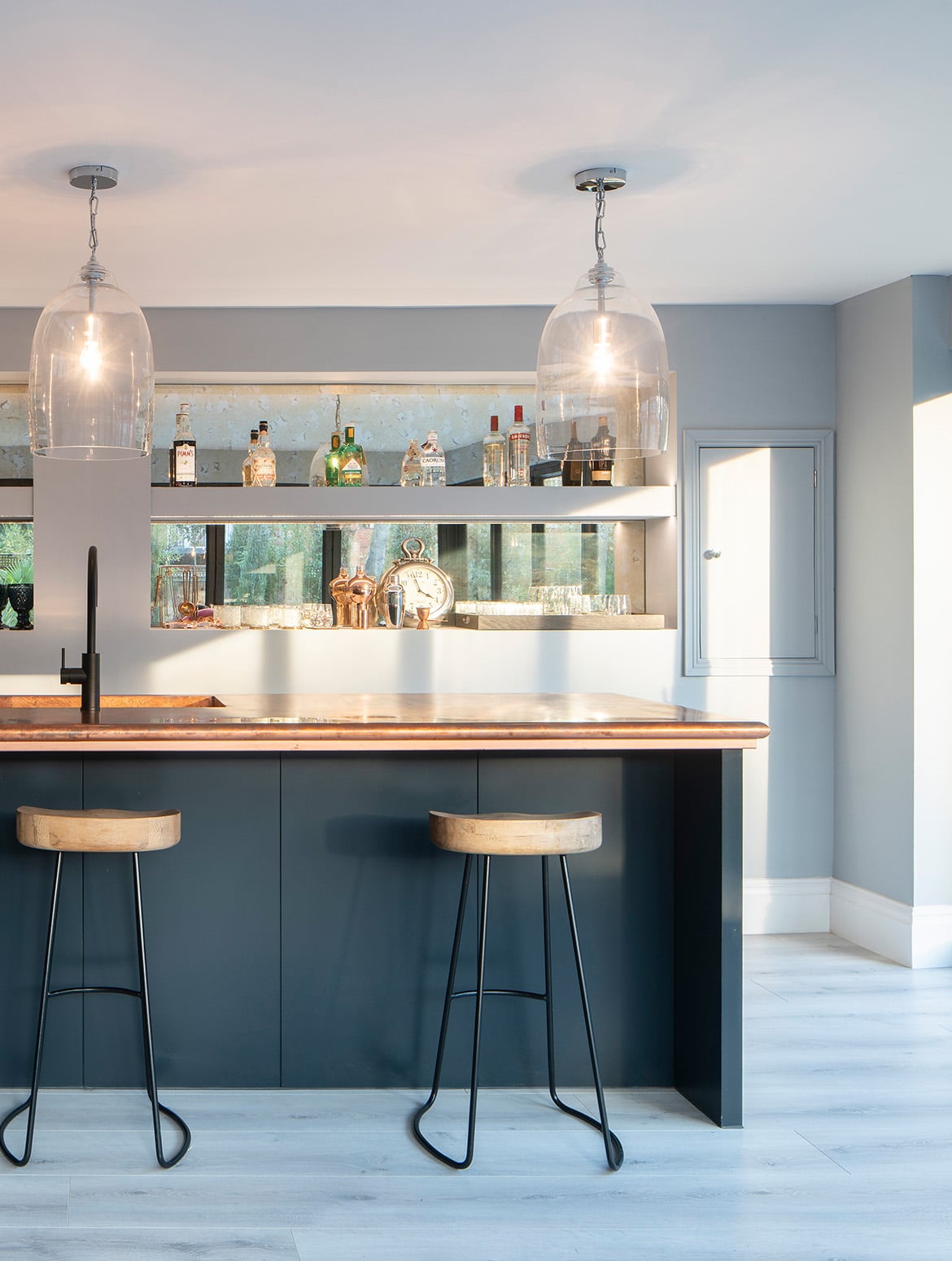A bespoke modern bar. The overhanging wooden surface of the bar offsets the black undersides, legs, and the two bar stools. The stools follow a similar pattern with wooden seats and black metal legs. A black Quooker tap stands before a sink, while two glass shades around bulbs above give the impression of upturned wine glasses. Behind the bar, two mirror-backed shelves have a selection of spirits and the glasses on them.  