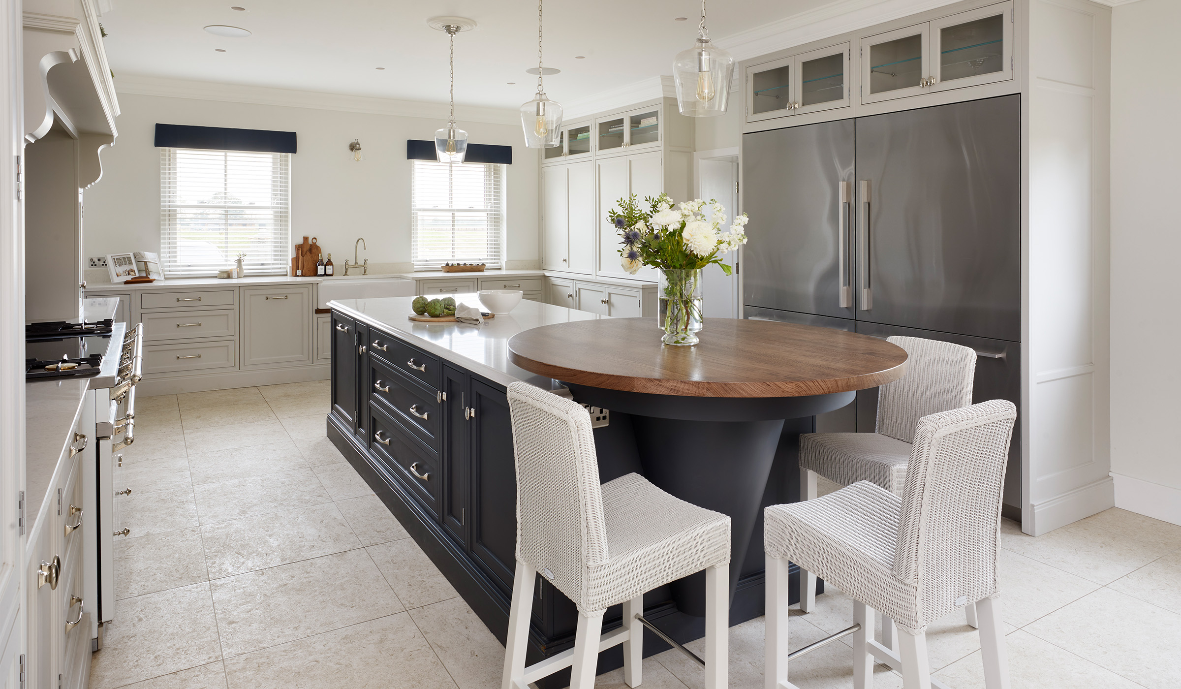 The Carr Farm Traditional Shaker Kitchen Project A traditional shaker kitchen in French Grey. The bespoke two-tiered central island in off-black Downpipe stands before a floor-to-ceiling row of cabinetry comprising cupboards, drawers, and Fisher & Paykel refrigeration. The wall opposite also has a mantel above a pastel blue Lacanche range cooker that stands proud to the rest of the bespoke cabinetry. Beyond the range cooker similar cabinetry and drawers line rest of the visible wall.