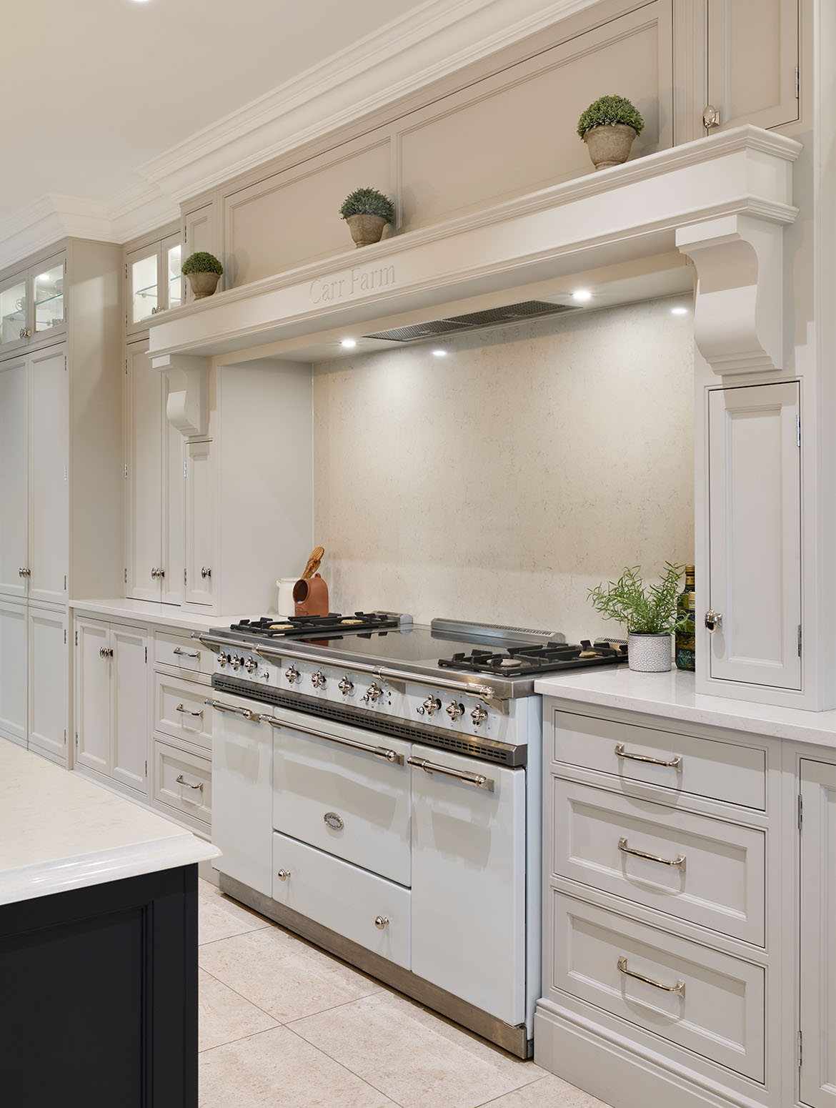 A traditional shaker kitchen in French Grey. The bespoke central island in off-black Downpipe stands before a floor-to-ceiling row of cabinetry comprising cupboards, drawers, and mantel above a pastel blue Lacanche range cooker. Beyond the range cooker similar cabinetry and drawers line rest of the visible wall. 