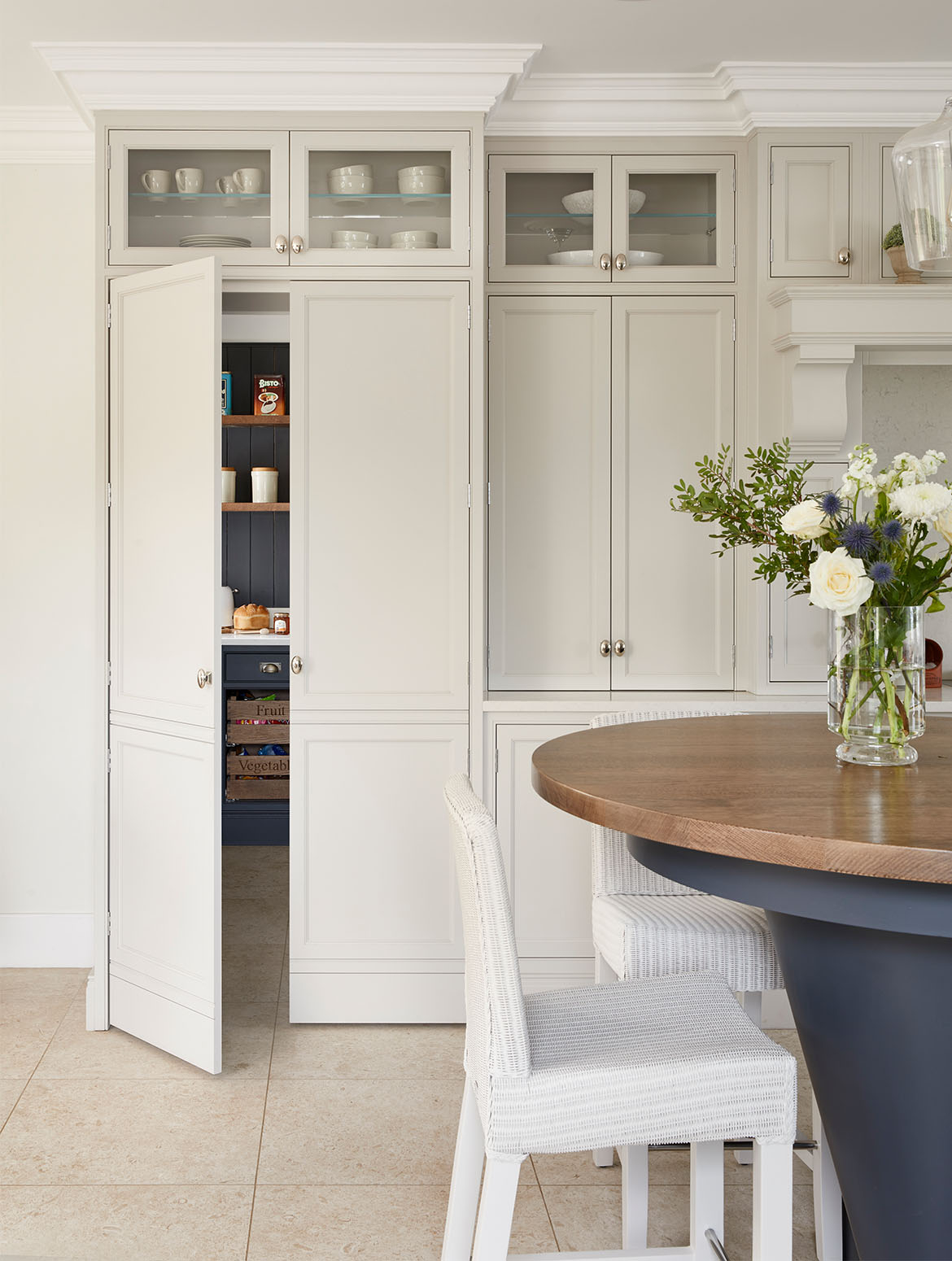 A traditional shaker kitchen in French Grey. The two-tiered central island in an off-black Downpipe contrasts the bespoke in-frame cabinetry and handles from Armac Martin’s Bournville collection. Two white wicker chairs stand below the second, higher tier. Beyond, two in frame floor-to-ceiling cabinets overlook the kitchen. The slight opening door of the left cabinet reveals it to be a hidden pantry with savoury items just visible inside. 