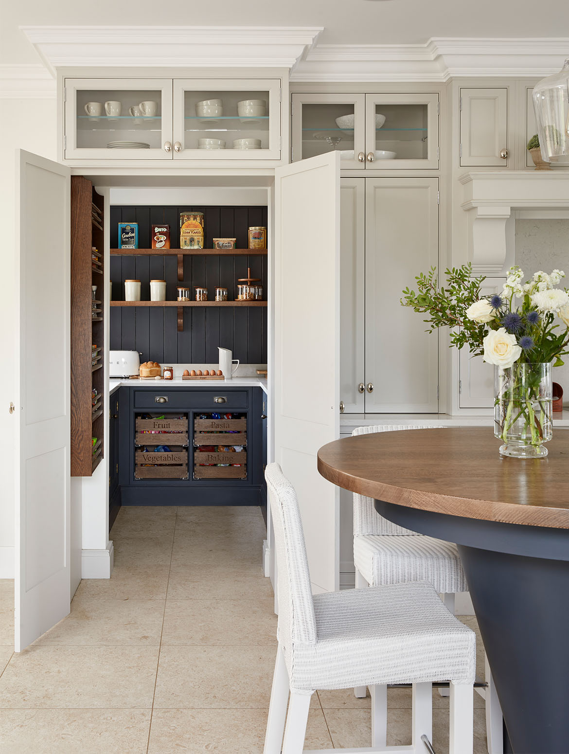 A traditional shaker kitchen in French Grey. The two-tiered central island in an off-black Downpipe contrasts the bespoke in-frame cabinetry and handles from Armac Martin’s Bournville collection. Two white wicker chairs stand below the second, higher tier. Beyond, two in frame floor-to-ceiling cabinets overlook the kitchen. The open doors of the left cabinet reveals it to be a hidden pantry with savoury items just visible inside. White worktops contrasts the handles and continued Downpipe cupboards and drawers. 