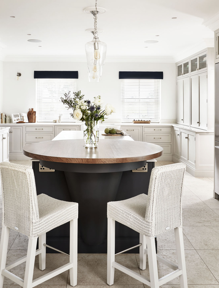 A traditional shaker kitchen in French Grey. The two-tiered central island in an off-black Downpipe contrasts the bespoke in-frame cabinetry. The left cabinetry is shaped around a white Lacanche range cooker. The mantel has Carr Farm engraved on it. To the right of the island, cabinetry surrounds a set of Fisher & Paykel fridge freezers. Along the back wall, bespoke pantry and the worktop stand below a pair of blinded windows.      