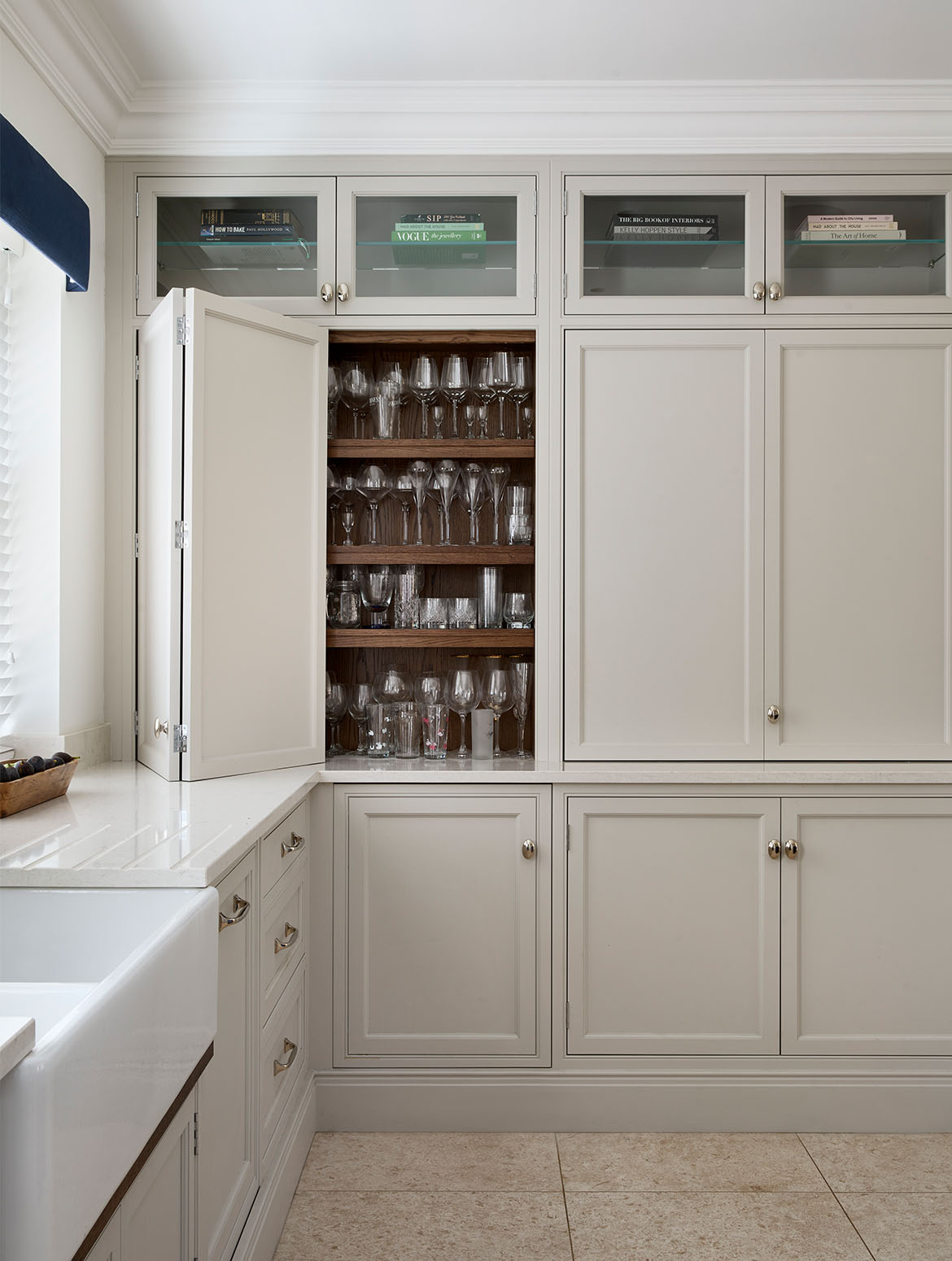 A traditional shaker kitchen in French Grey. The right-angled cabinetry hugs the corner of room. The left side has a white worktop with bespoke cupboards below with Armac Martin handles. The open cupboard of the floor-to-ceiling cabinetry reveals four shelves of different sized glasses. 