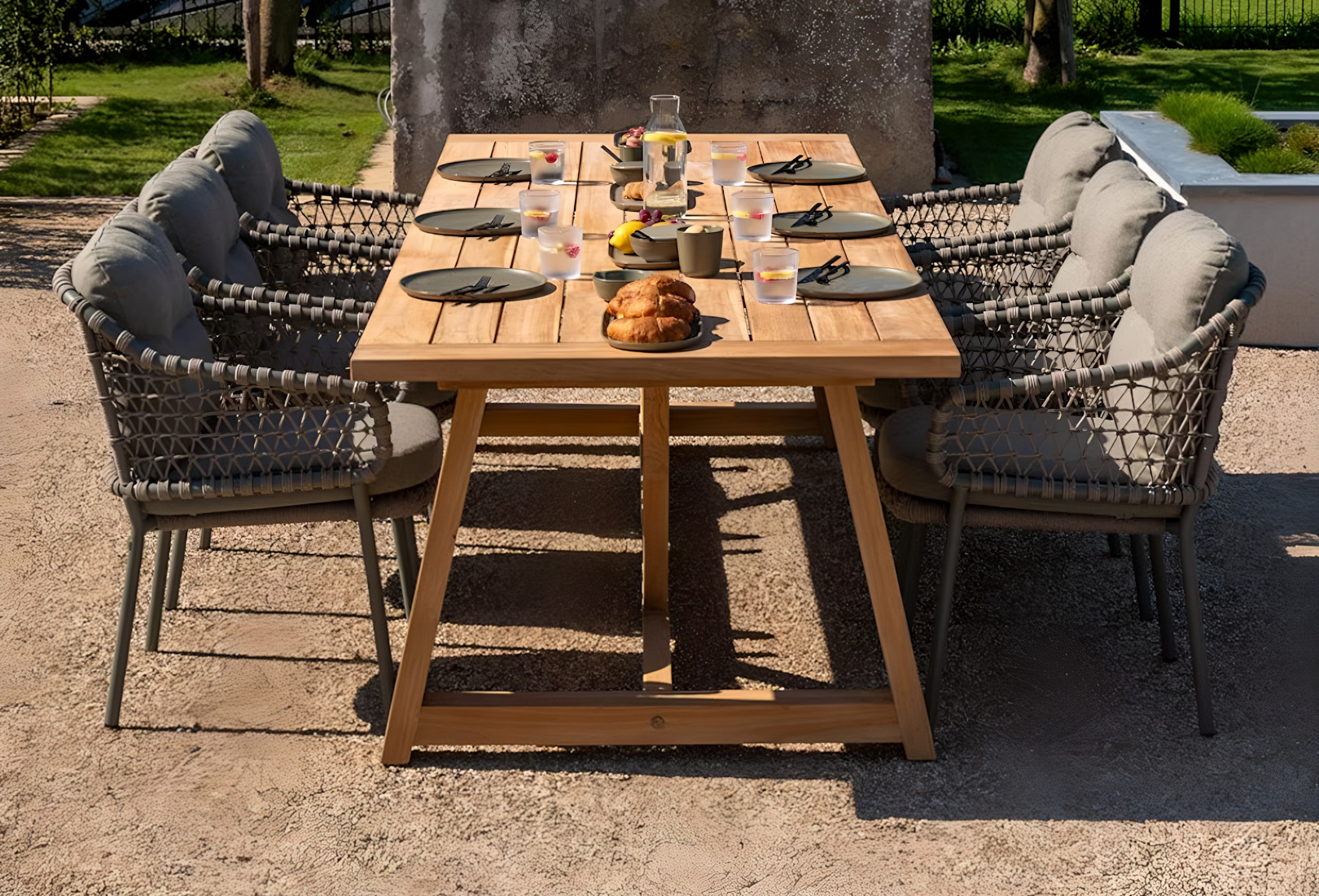 An image of 4 Season Outdoor’s Jura dining set. The dining set comprises three chairs either side of a long beige wooden table. The olive green chairs have knotted, rope-wrapped sides to them and olive green seat cushions. The teak dining table contrasts the dirt floor well and is set for an outdoor breakfast with fruit, croissants and a jug of water in the middle of the table, along with place settings for each seat. 
