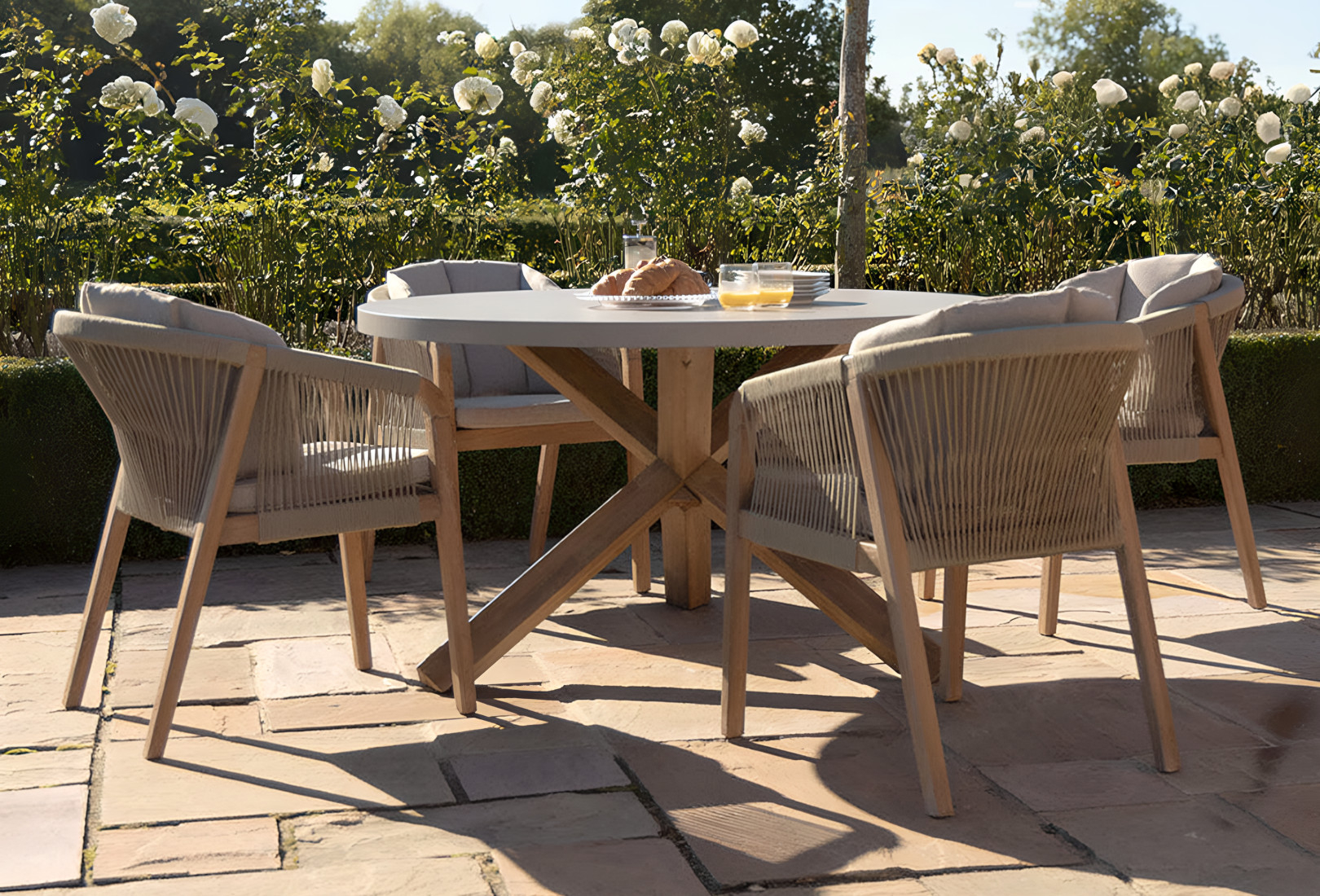 An image of Maze Living’s Martinique Rope Weave 4 Seat Round Dining Seat. The light brown antique finish of the four chairs contrast the acacia wooden frames and the ample seat cushions. The matching cross-legged dining table has plates for croissants and a dish with the pastry on it next to two glasses with a dash of orange juice in them. The patio has a bush-lined perimeter with white rose bushes behind. 