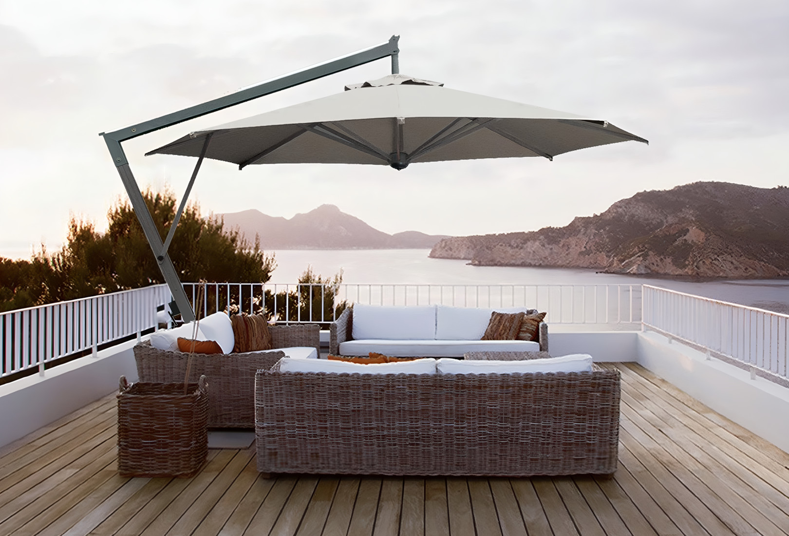 An image of Scolaro Parasol’s Galileo Maxi Dark parasol. The cantilever parasol oversees a wicker three sofa lounge set with white cushions. The wooden balcony they’re positioned on oversees a lake at sundown. 