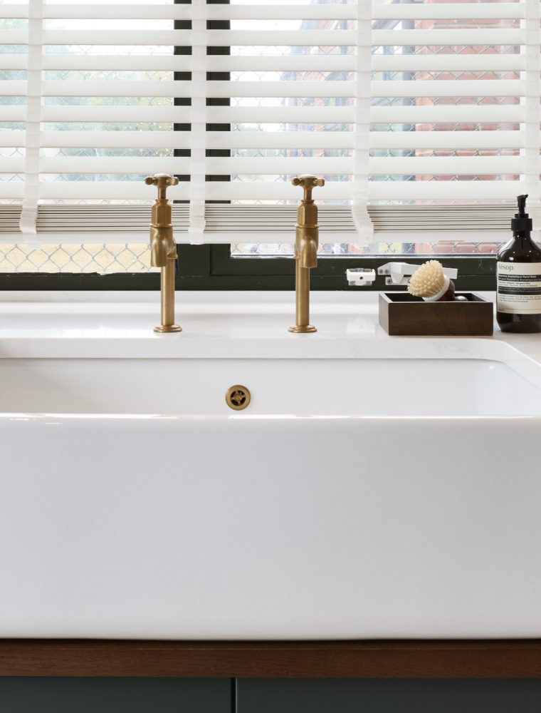 A bespoke bootroom in Studio Green. A close in view of the Belfast sink and burnish taps. Next to the cold tap, a small tray with a brush and hand soap stands opposite a small ornamental glass vase. The bespoke cabinetry below the sink contrasts well with the white worktop above. 