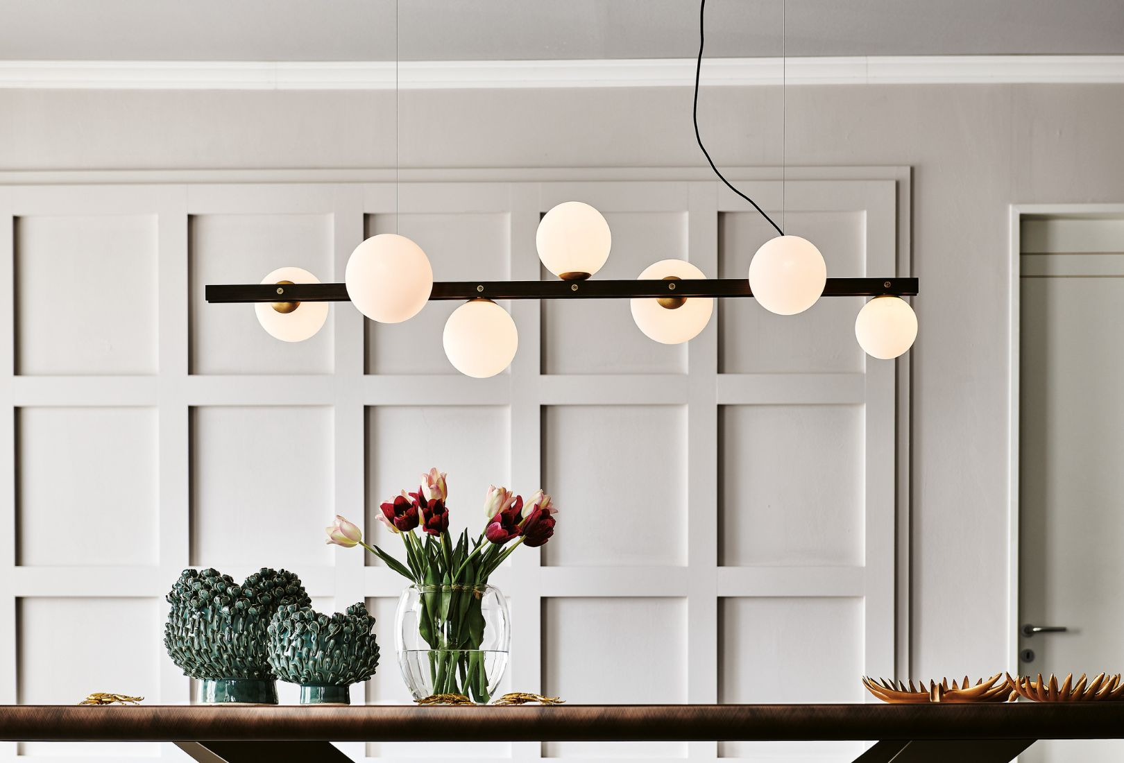 A wood-panelled wall in white foregrounds Cattelan Italia’s Planeta light fixture. At intervals, the white glass spheres line the bar fixture suspended by two thin wires either side of it. Underneath, the table has two ceramic and glass vases with succulents in them to left and to the right two gold painted ceramic trays that look like flowers laid on their back. 