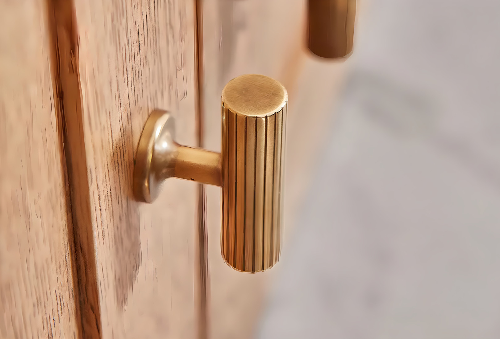 A close in view of a T-handle from Armac Martin. The antique brass handle has fluted edging around it and attaches to wooden door that has a light texture. 