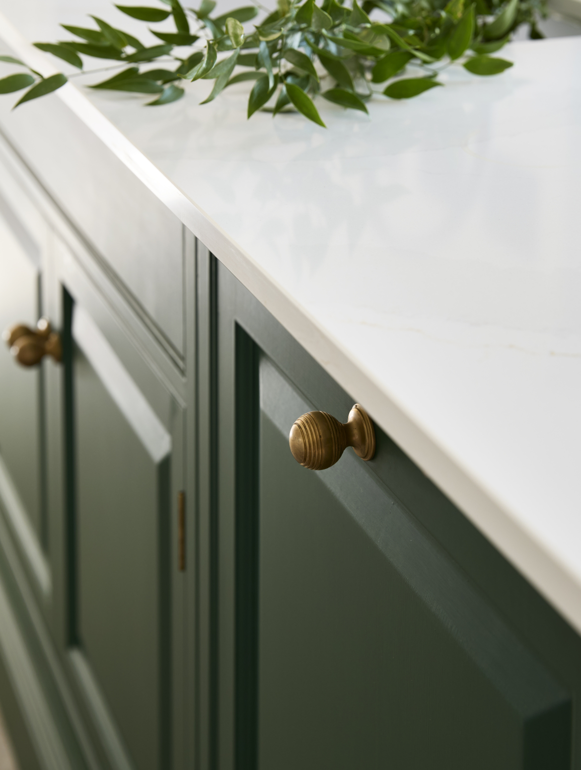 A traditional shaker kitchen with a two-tier island and green bespoke cabinetry. The white island top has the branch of bay laid across it while below the Perrin and Rowe handles on the cupboards and drawers below stand in contrast to the green cabinetry. 
