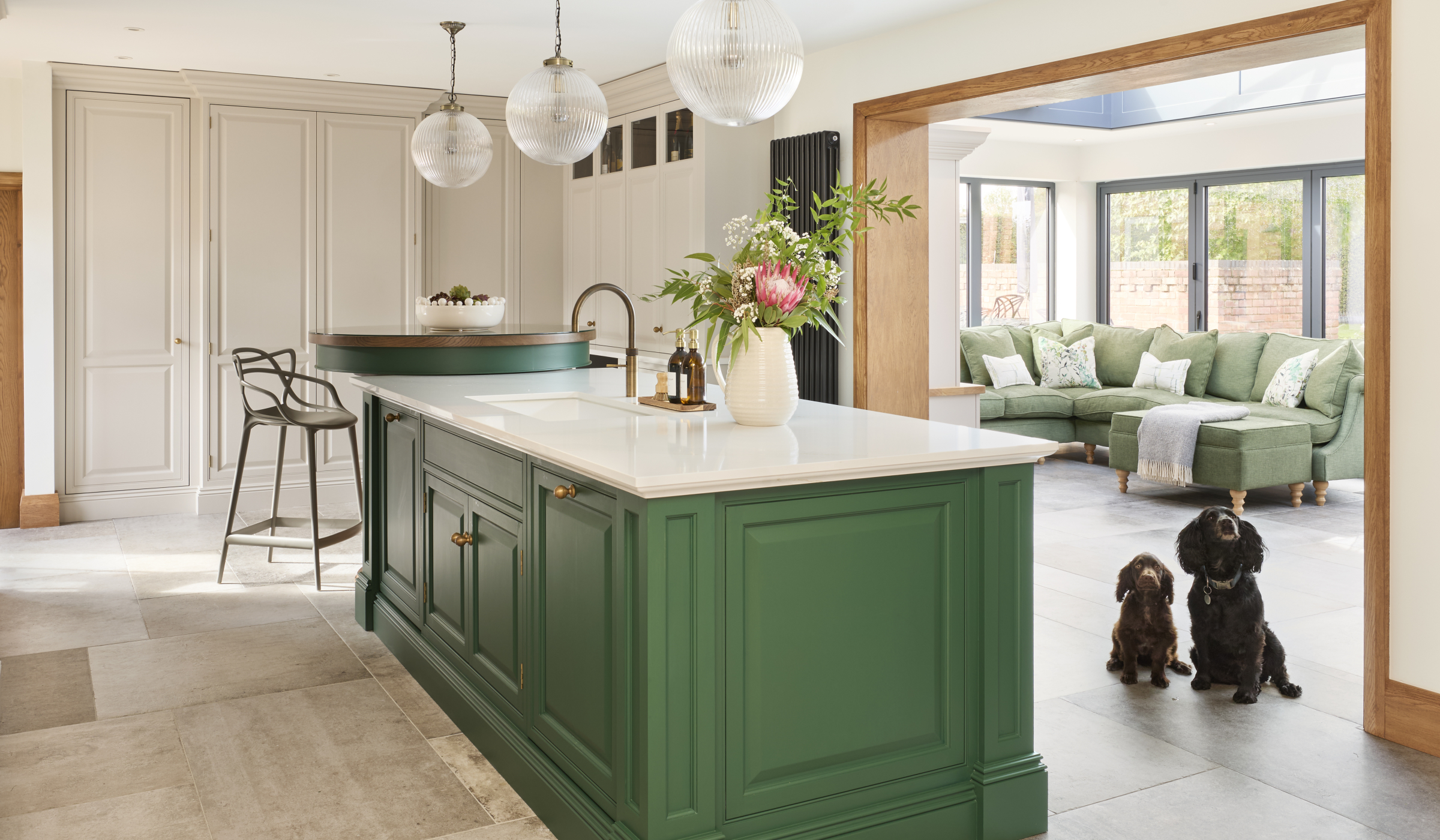 The Brunswick Traditional Shaker Kitchen Project A traditional shaker kitchen with a two tier island with green bespoke cabinetry. Two Cocker spaniels sit beside the bespoke island fitted with a Quooker tap and Shaws sink and has a raised second tier accommodated a two metal high chairs. The spaniels sit in the walkthrough to the open and spacious living area—complete with a green L-shaped sofa.