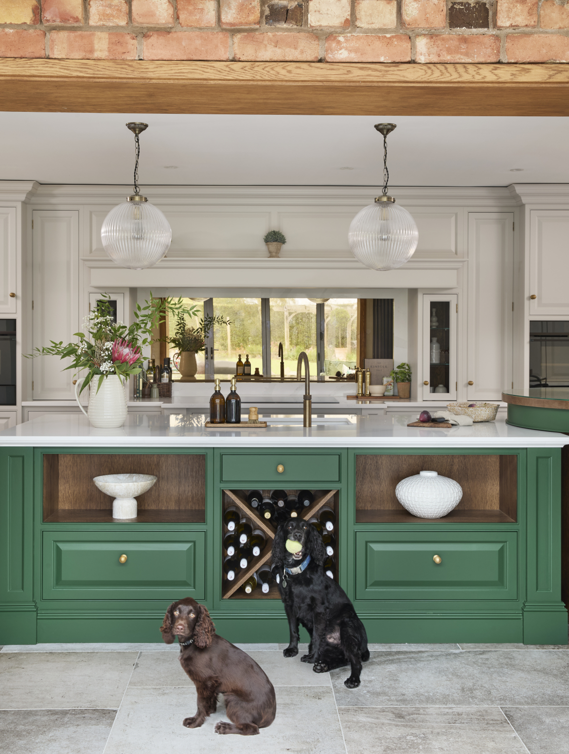 A traditional shaker kitchen with a two-tier island and green bespoke cabinetry. Two spaniels sit in front the island’s discreet wine cabinet—the older of the two has a tennis ball in its mouth. Behind them there is a Quooker tap central to the island as well as a ceramic vase of flowers. Behind the island the bespoke French Grey cabinetry oversees the kitchen. 