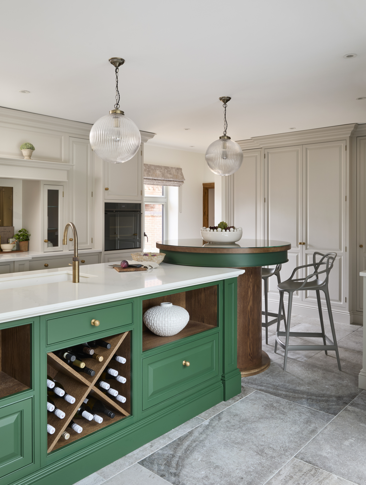 A traditional shaker kitchen with a two-tier island and green bespoke cabinetry. The Brunswick green bespoke island cabinetry shows a discreet wine cabinet also niches where a two ceramic vase sit. The taller tier of the island sits behind surrounded by two metal high chairs.