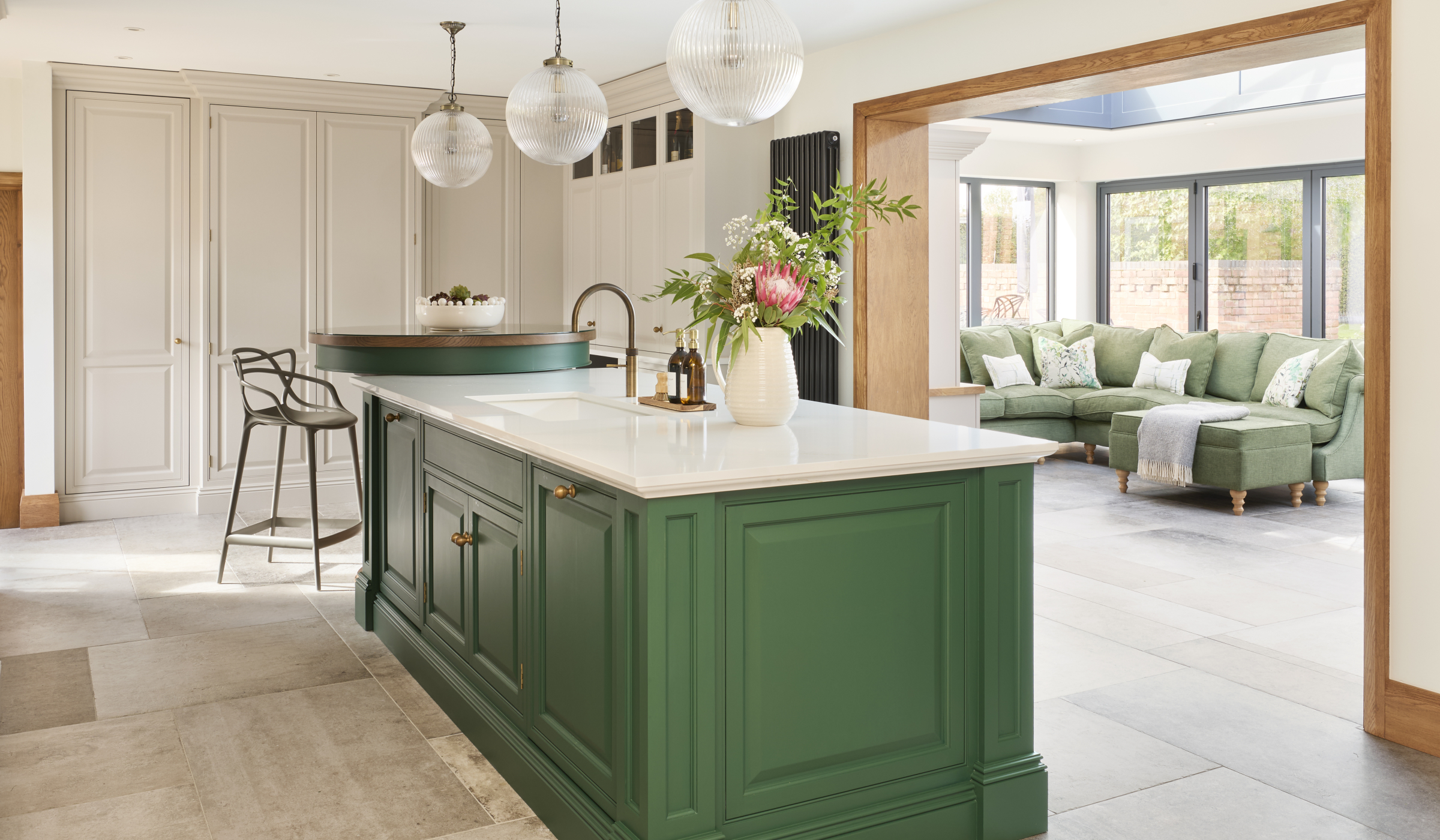 The Brunswick Traditional Shaker Kitchen Project A traditional shaker kitchen with a two tier island with green bespoke cabinetry. The bespoke island fitted with a Quooker tap and Shaws sink, has a raised second tier accommodated a two metal high chairs. There is a walkthrough to the open and spacious living area—complete with a green L-shaped sofa.