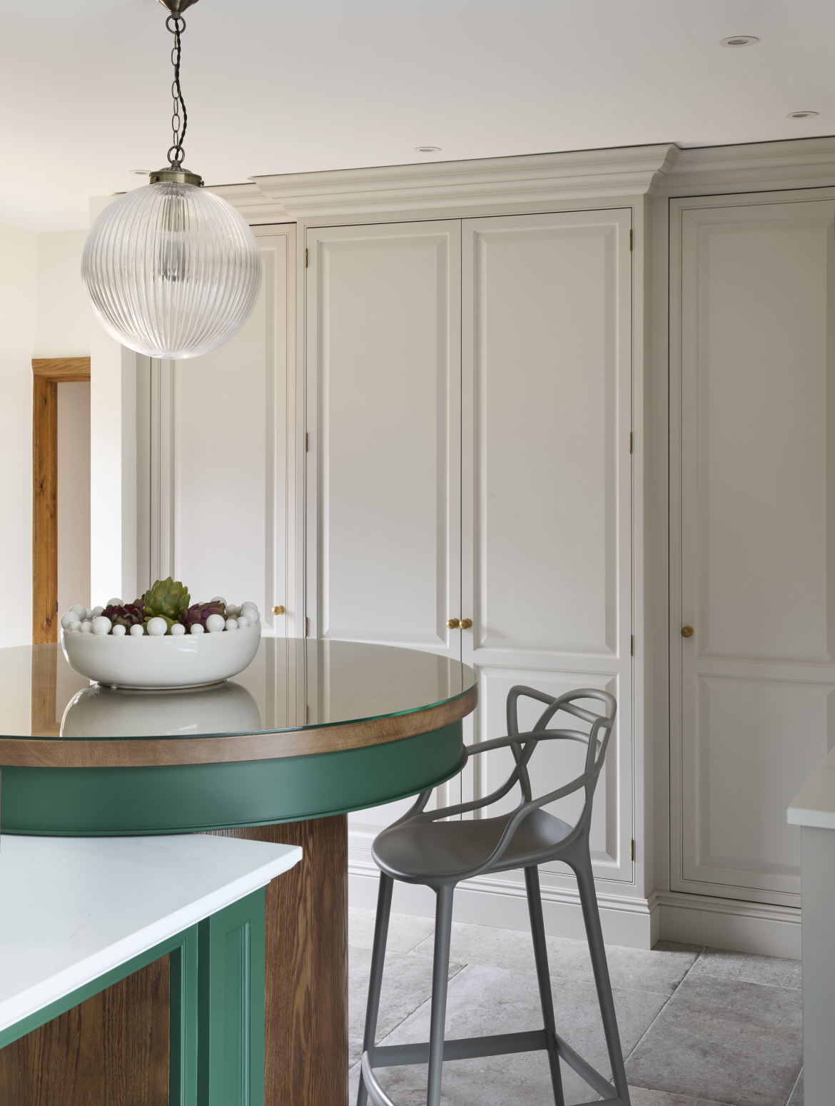 A traditional shaker kitchen with a two-tier island with green cabinetry. The second, and taller tier, is an even circle with a metal high chair positioned underneath it. Behind floor to ceiling bespoke cabinets in French Grey offset the Brunswick green of the island.
