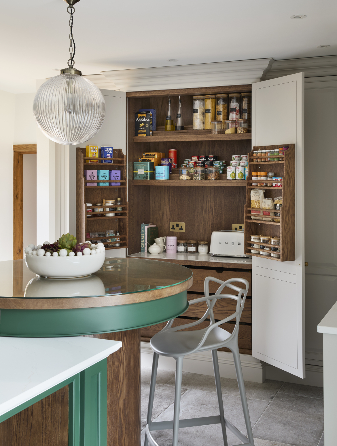 A traditional shaker kitchen with a green two-tier island with bespoke cabinetry. The second, and taller, island tier is an even circle with a metal high chair underneath it and it foregrounds floor to ceiling bespoke pantry in French Grey which offsets the Brunswick green of the island. This pantry has both doors open, showing a variety of ambient savoury foods and hot beverage essentials.
