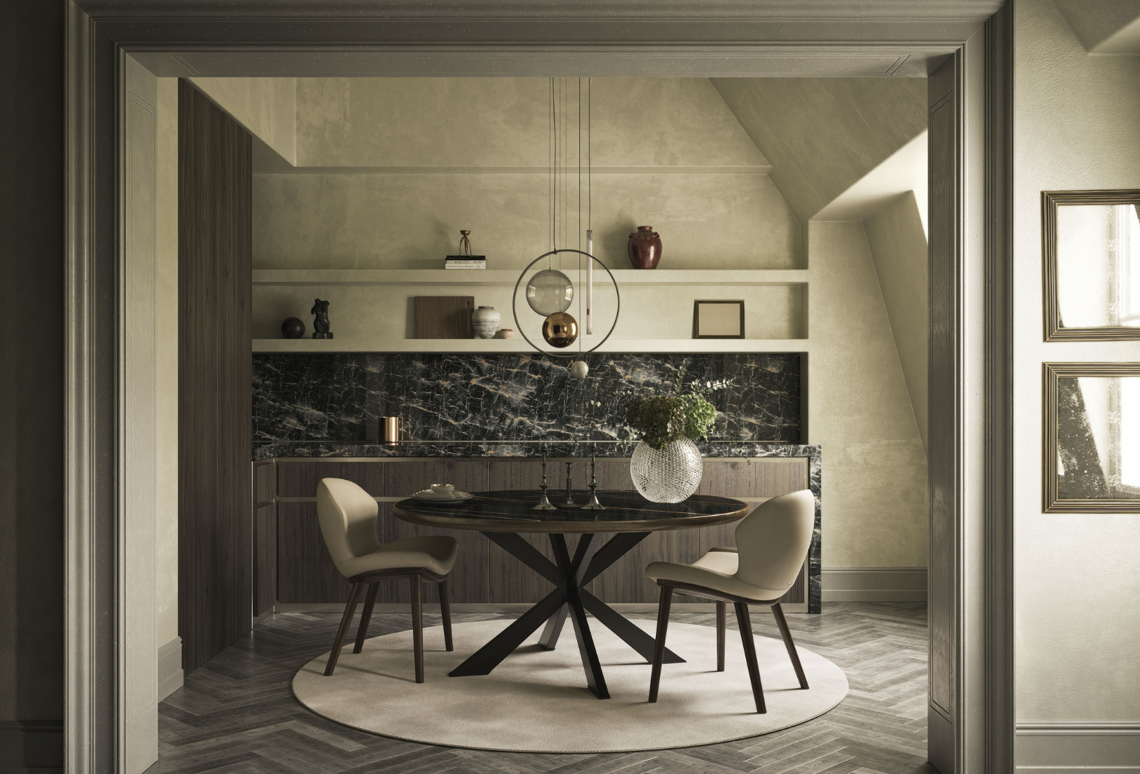 A modern area framed by a square archway. The circular table has two upholstered cream chairs around it on angles opposite each other. On the table, three candle sticks and a large spherical glass vase stand beneath a pendant light fixture with a selection of orb, icicle, and hoop shades and mounts. The console with a blackish white marble finish beyond runs along most of the back wall. On the two shelves above, a selection of ceramic vases and jugs, picture frames and statuettes catch shadows from the window out of view to the right. Two frame one above the other on the archway wall also reflect light from a second window out of view.