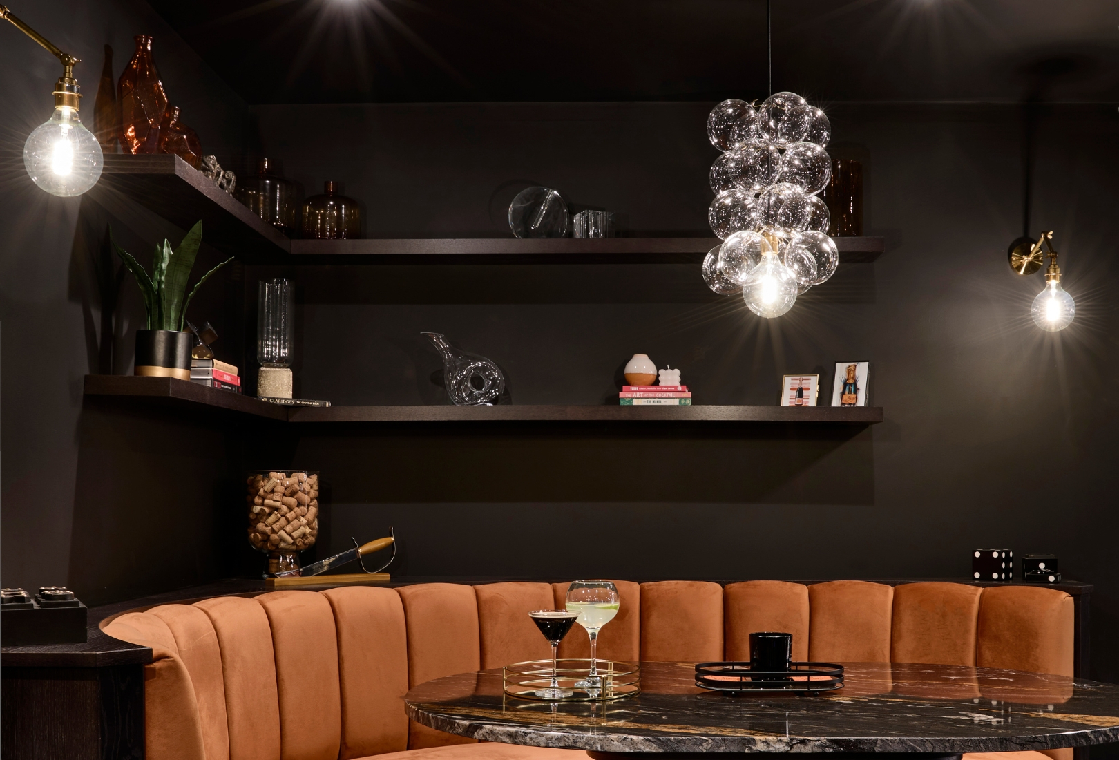 A bespoke bar area in onyx. The orange velvet booth set into the bespoke cabinetry melds easily with the Off-Black walls around it. The Orinoco Granite table reflects light from the chandelier above along with the two cocktails on the table. The shelving above moulds around the corner and has a selection of books, memorabilia, ceramic pots, and plants. 