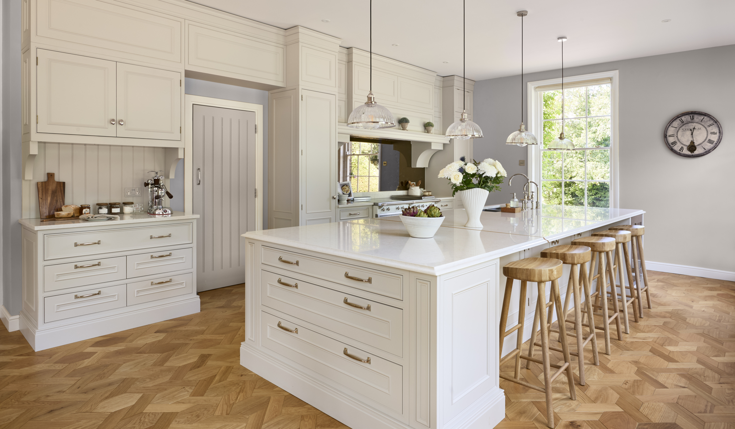 The Rose Garden Traditional Shaker Kitchen Project A traditional cream shaker kitchen with a central island and bespoke cabinetry. Hanging lights overlook the island while wooden stools sit underneath an exposed lip in worktop. On the other side cabinets and worktop sit flush to one side of a doorway and on the other a white Lacanche range cooker stands proud to the other cabinet beside and above. An end corner of the island also as two Quooker taps.