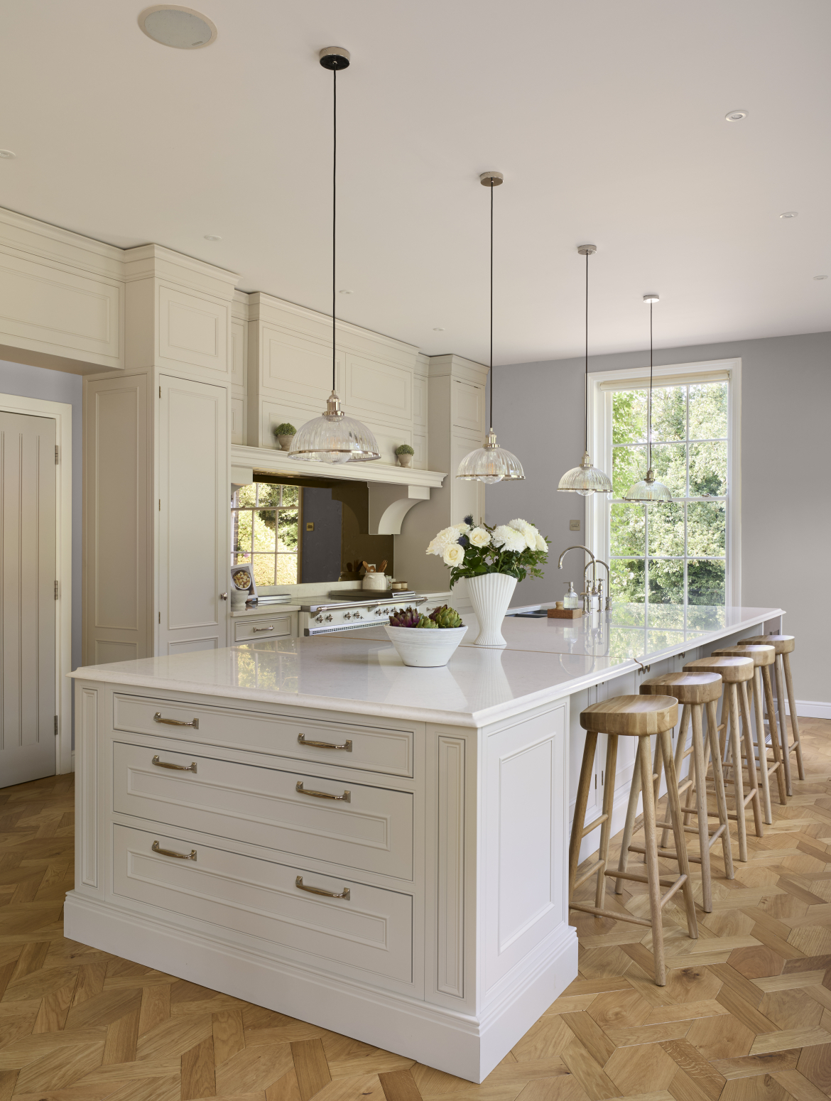 A traditional cream shaker kitchen with a central island and bespoke cabinetry. The island has end-drawers and stools that slide underneath the island’s edge. While on top there are two Quooker taps opposite a white Lacanche range cooker. 