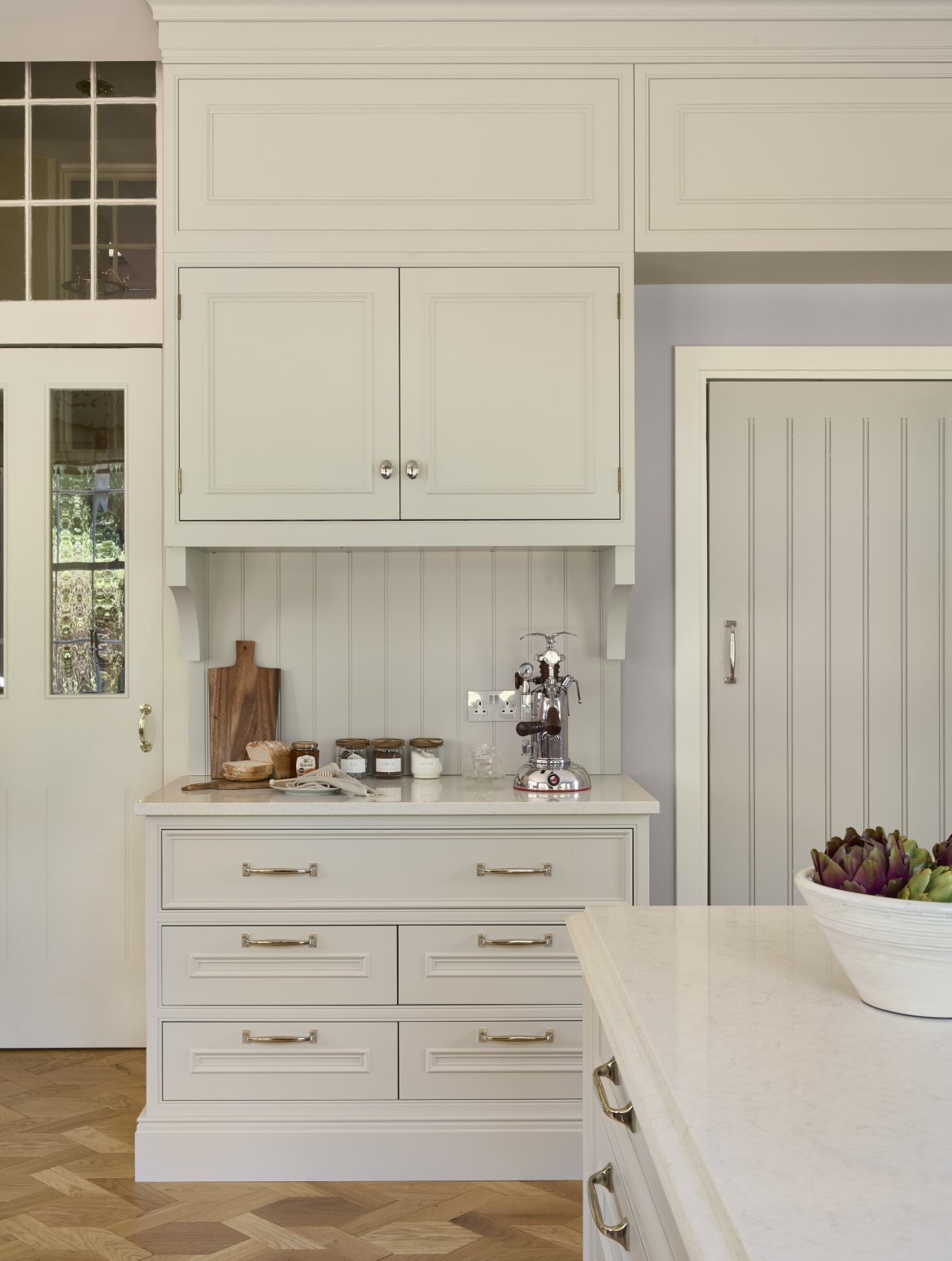 A traditional cream shaker kitchen with bespoke cabinetry and island. The worktop, set between two, doorways, holds La Pavoni coffee machine and the necesssary items for a warm beverage: tea, coffee, and sugar.  