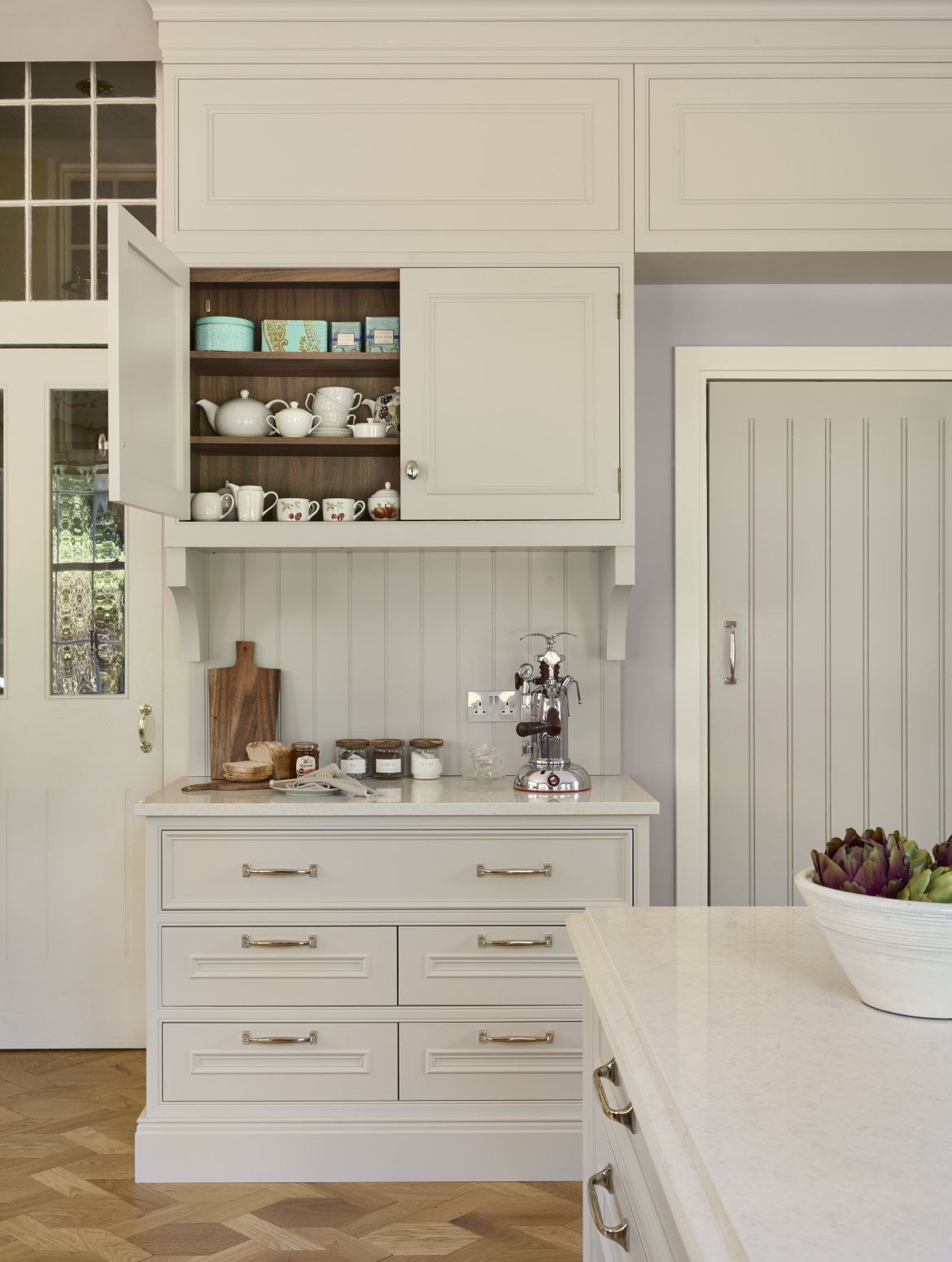 A traditional cream shaker kitchen with bespoke cabinetry and island. The worktop, set between two, doorways, holds La Pavoni coffee machine and the necessary items for a warm beverage: tea, coffee, and sugar. While above, the open cupboard has an assortment of teapots, teacups, and Fortnum and Mason tea.