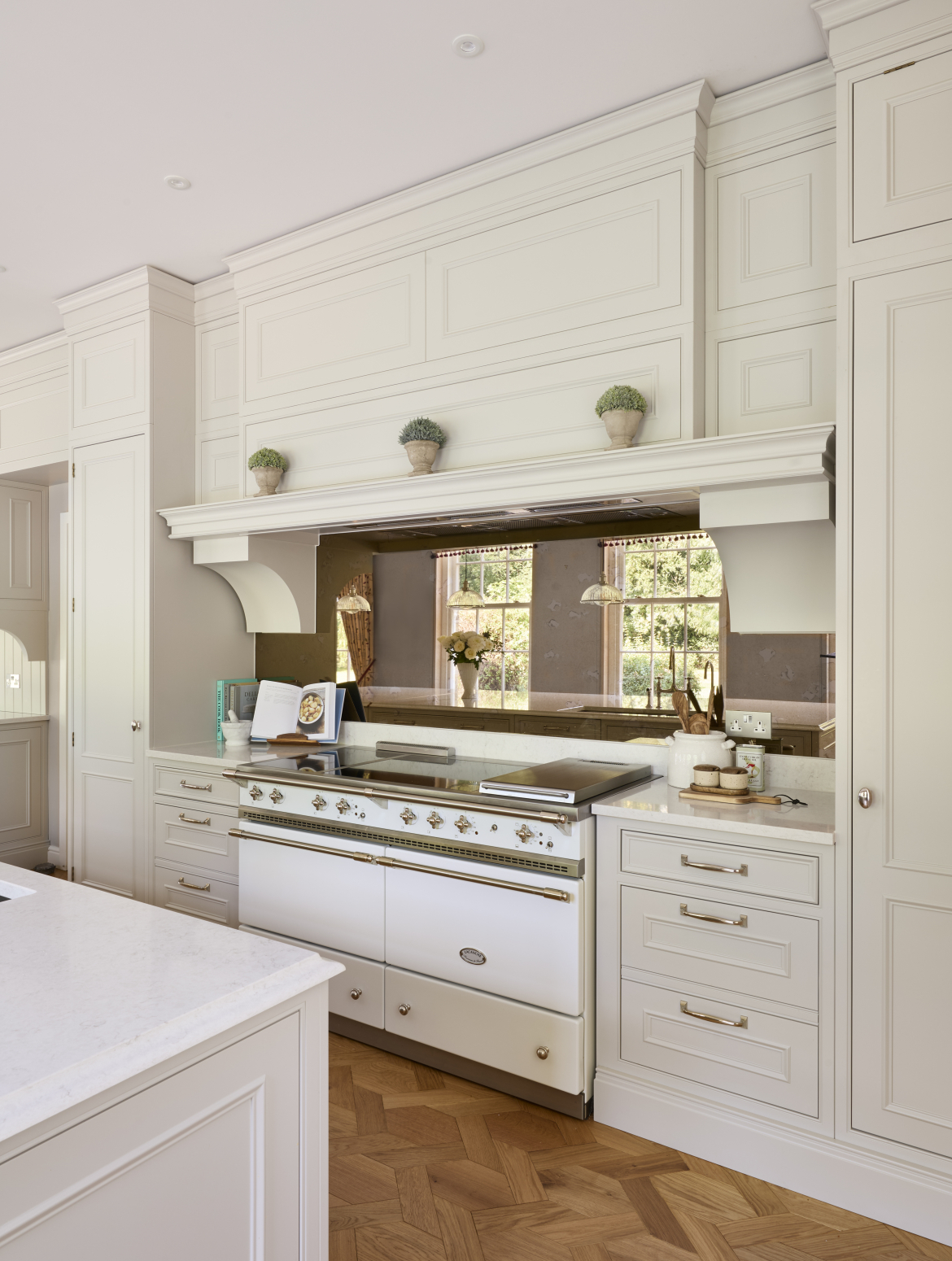 A traditional cream shaker kitchen with bespoke cabinetry around a white Lacanche range cooker. On one side of it, there are utensils and seasoning and on the other there are cook books. 