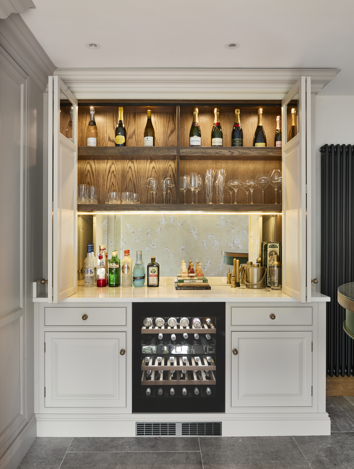 A traditional shaker kitchen with bespoke cabinetry. A floor-to-ceiling bespoke cabinet housing a bar contains a variety of spirits in different coloured bottles and champagne. An under-counter wine fridge sits central to the lower half of the cabinet. Above the bottles, two separate shelves contain champagne and a selection of different glasses. 