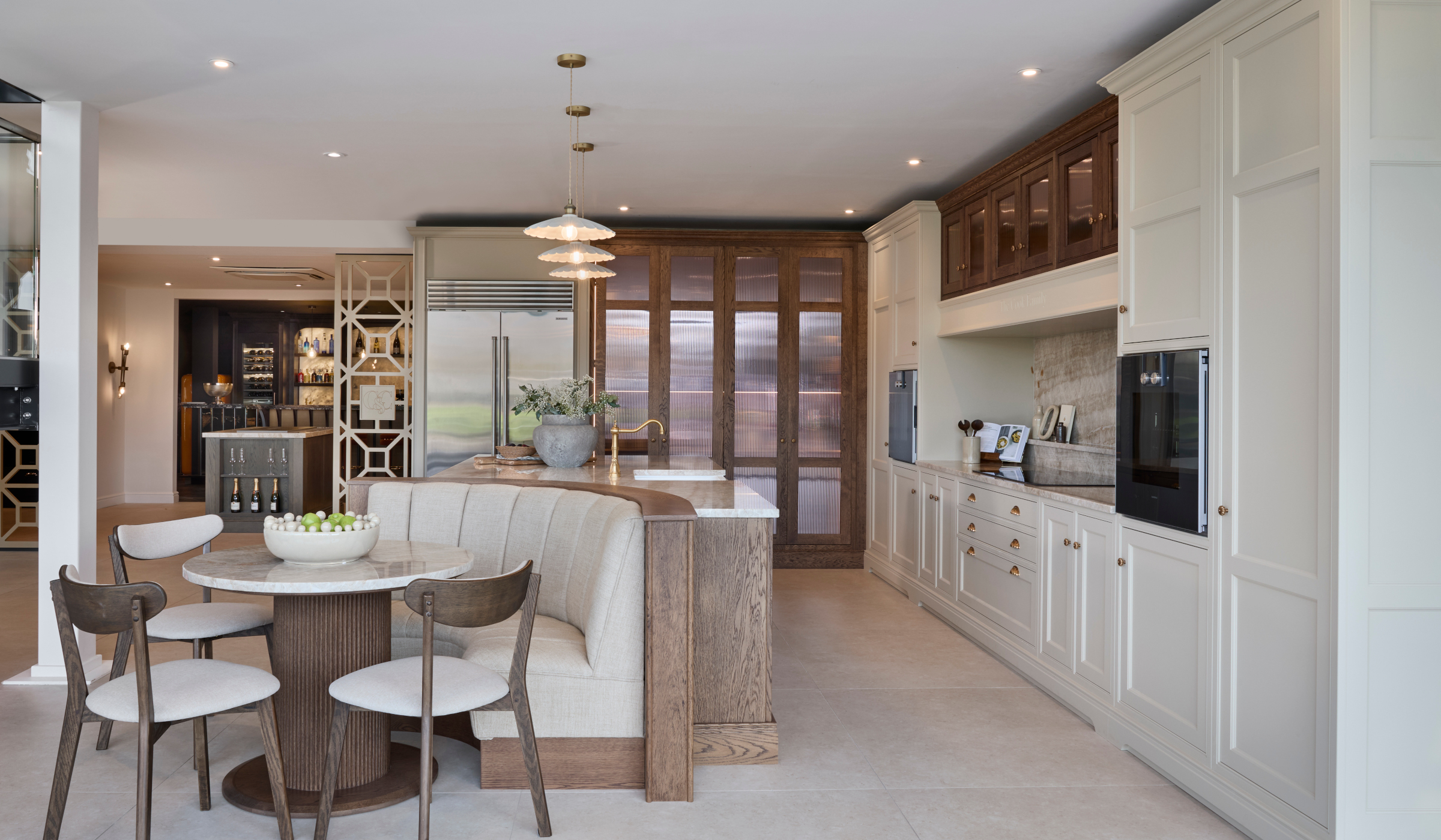 The Cooks Family Traditional Shaker Kitchen Project A traditional shaker kitchen in French Grey. A bespoke island with a seating area embedded within its curved edge stands centrally with three upholstered chairs around it. The Taj Mahal stone stands out from the Stony Ground colour of the bespoke cabinetry beyond and adjacent. To the right side of the island stands bespoke cabinetry surrounding the mantel. Two Gaggenau ovens stand proud on each side. Beyond the island, the floor-to-ceiling pantry and Sub-Zero fridge freezer stand next to each other. Armac Martin handles around the kitchen provide a further contrast to the Taj Mahal marble.
