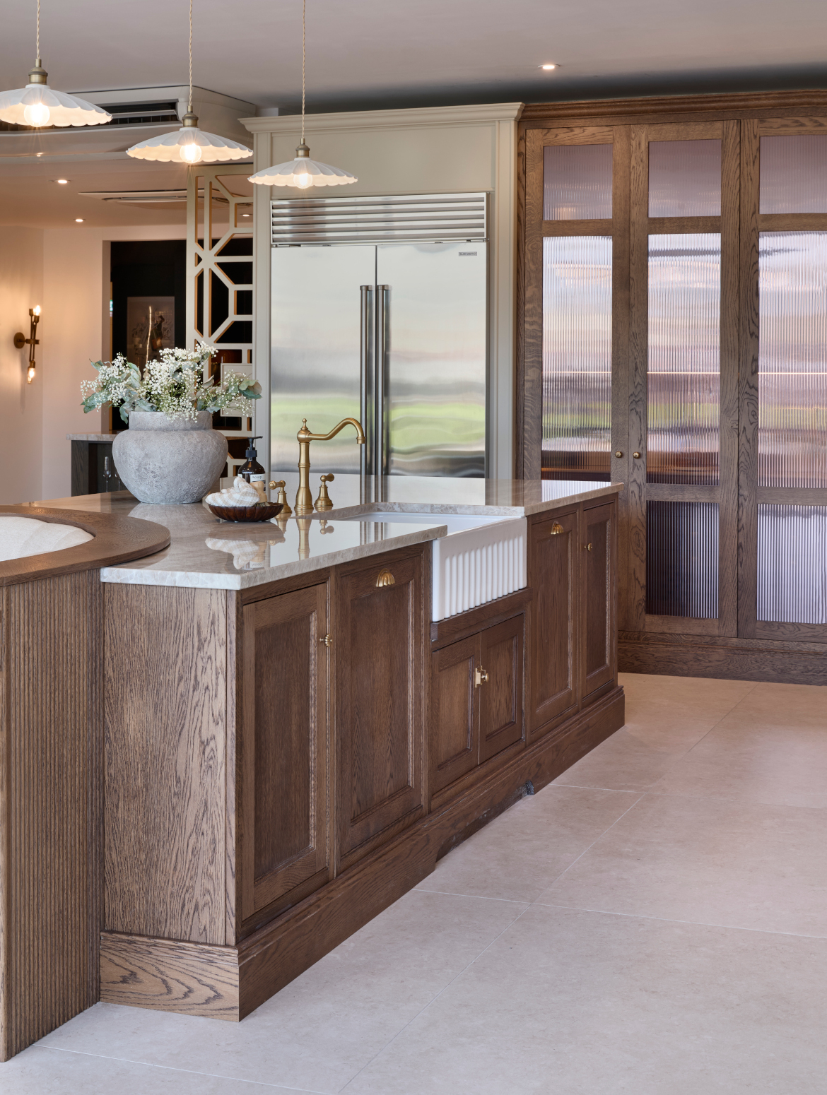 A traditional shaker kitchen in French Grey. A bespoke island with a seating area embedded within its curved edge stands centrally. The island’s Taj Mahal stone stands out from the Stony Ground colour of the bespoke cabinetry beyond. Beyond the island, the floor-to-ceiling pantry and Sub-Zero fridge freezer stand next to each other. Armac Martin handles around the kitchen provide a further contrast to the Taj Mahal marble.
