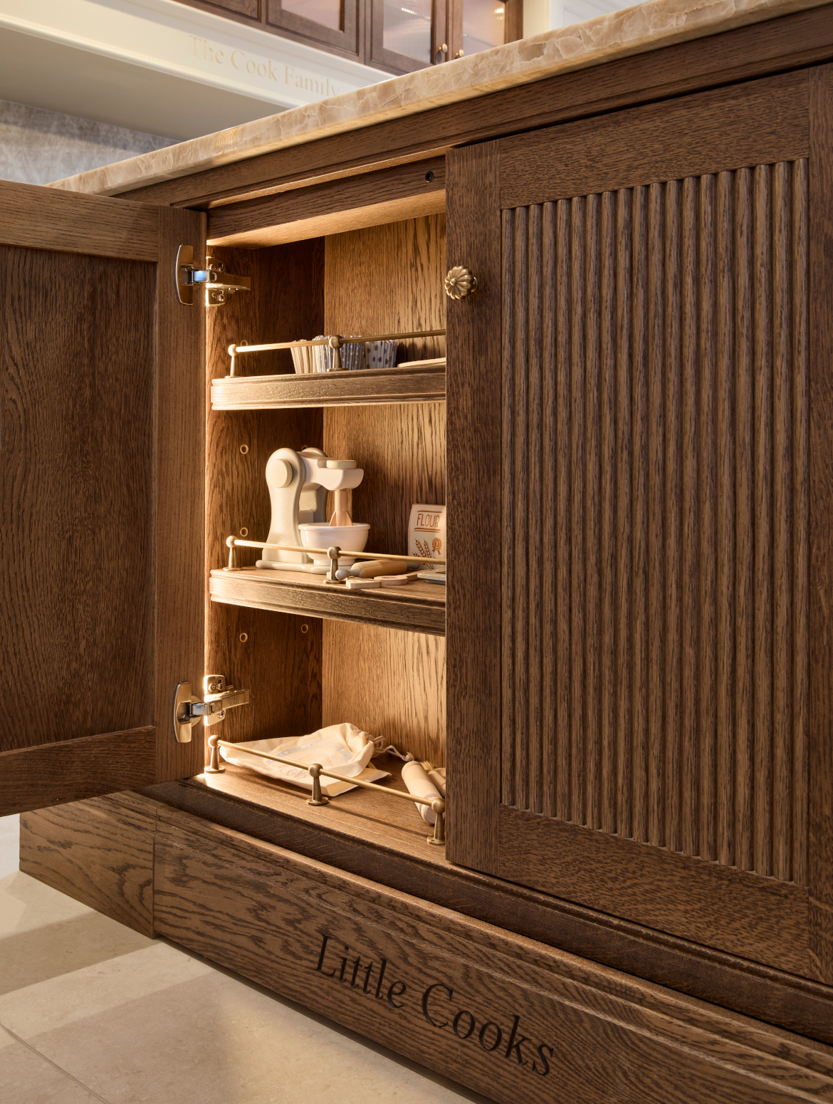 A traditional shaker kitchen in French Grey. A bespoke central island cupboard handled in Armac Martin. The open door reveals three shelves with wooden toys of kitchen utensils. The mantel below is engraved with: Little Cooks. 