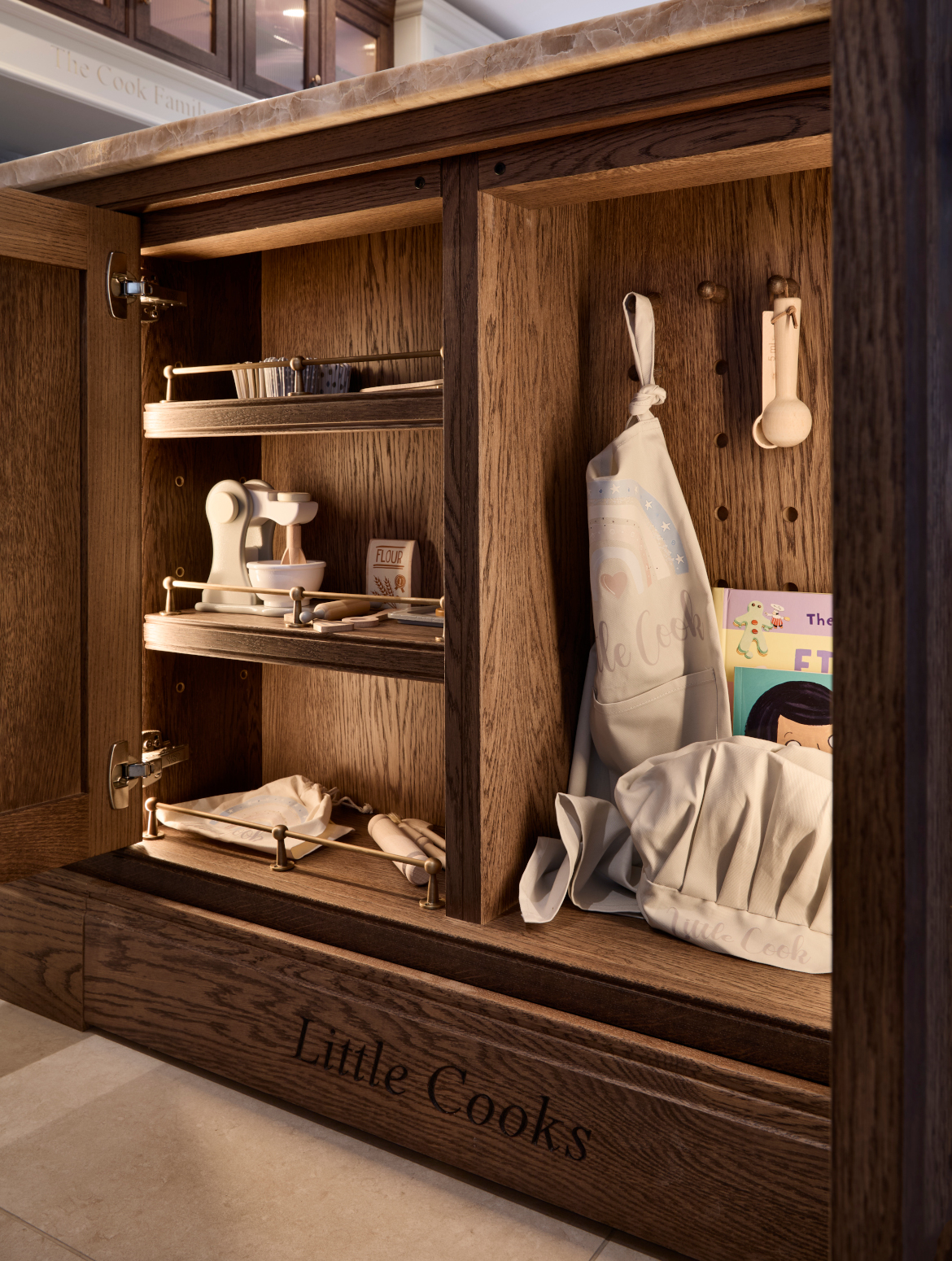 A traditional shaker kitchen in French Grey. A bespoke central island cupboard handled in Armac Martin. The open door reveals three shelves with wooden toys of kitchen utensils. The mantel below is engraved with: Little Cooks. The second open cupboard door has hooks for a child-sized apron and hat. 