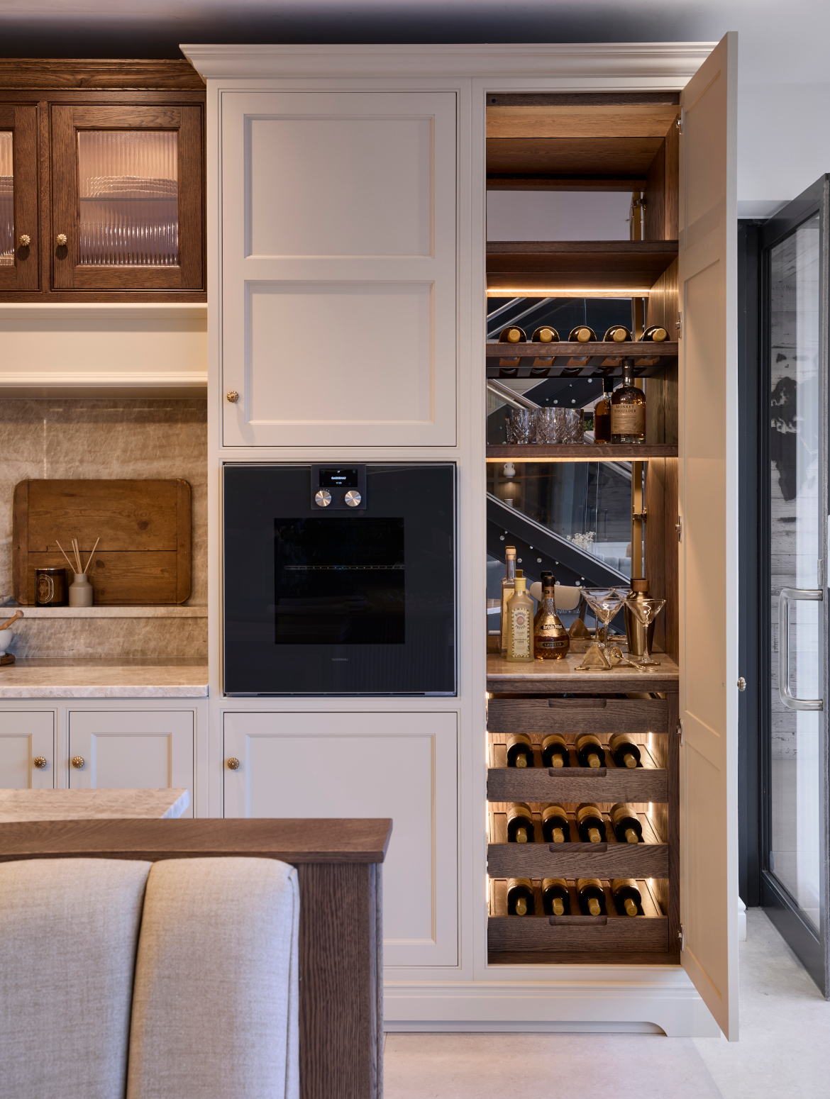 A traditional shaker kitchen in French Grey. The bespoke seating embedded in the central island is just visible before the bespoke cabinetry surrounding the hob. The floor-to-ceiling cabinet has a Gaggenau oven built into one side and the other reveals a mixology section. 