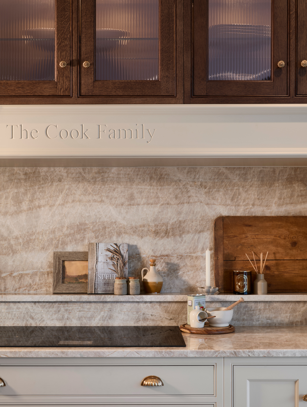A traditional shaker kitchen in French Grey. The mantel over the built-in induction hob has The Cook Family inscribed on it. Above the stained wood cabinetry with Armac Martin handles with fluted glass alludes to the crockery inside. The accoutrements for cooking such as cooking oils, chopping boards, and salt stand on the worktop and shelf behind the hob. Drawers beneath the hob worktop can just be seen. 