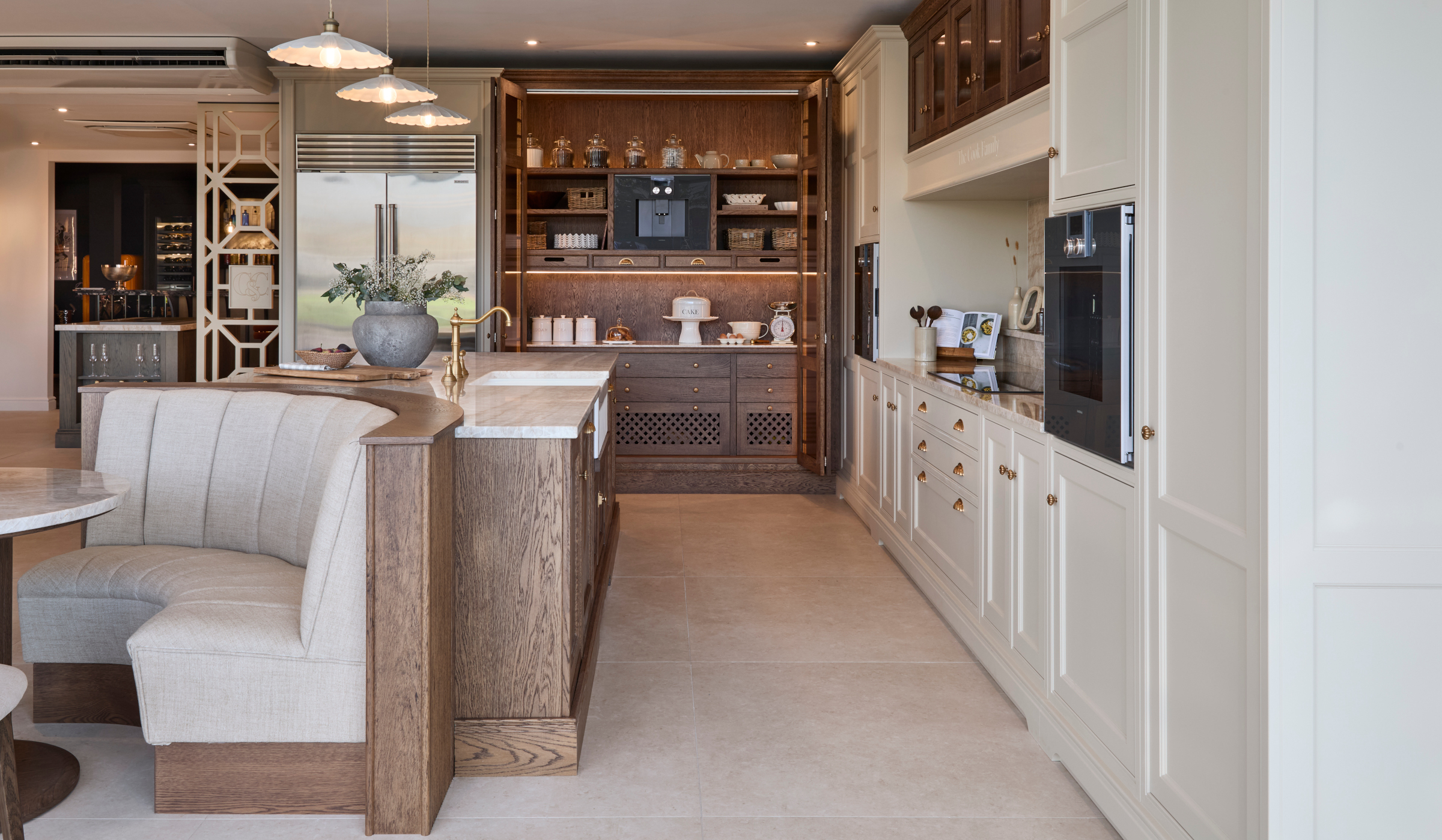 The Cooks Family Traditional Shaker Kitchen Project A traditional shaker kitchen in French Grey. A bespoke island with a seating area embedded within its curved edge stands centrally. The Taj Mahal stone stands out from the Stony Ground colour of the bespoke cabinetry beyond and adjacent. To the right side of the island stands bespoke cabinetry surrounding the mantel. Two Gaggenau ovens stand proud on each side. Beyond the island, the open pantry reveals Armac Martin-handled drawers, a baking station and a Gaggenau coffee machine. A Sub-Zero fridge freezer stands alongside the pantry.
