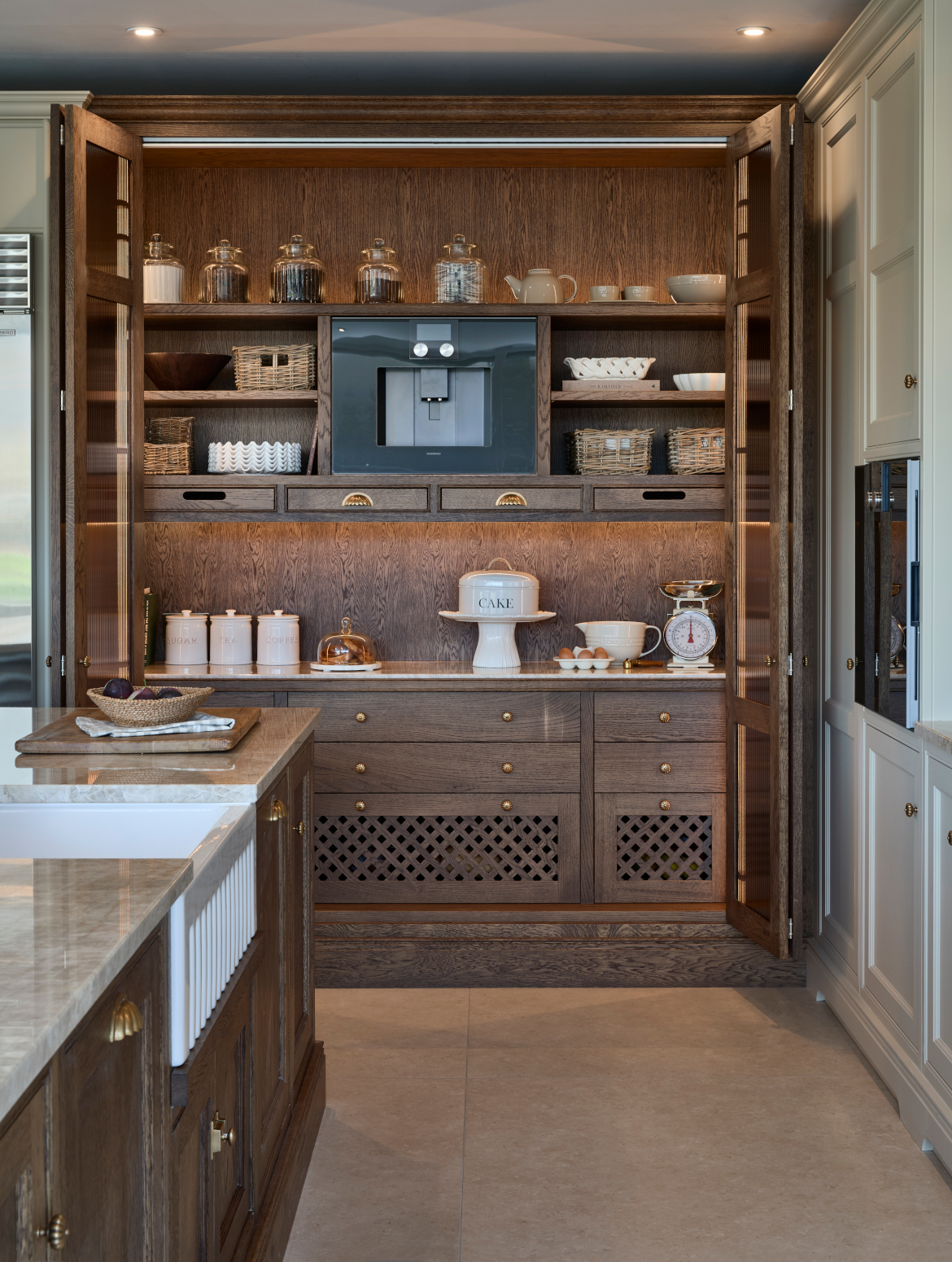 A traditional shaker kitchen in French Grey. A bespoke island with Taj Mahal stone stands out from the Stony Ground colour of the bespoke cabinetry beyond and adjacent. Beyond the island, the floor-to-ceiling pantry and Sub-Zero fridge freezer stand next to each other. Armac Martin handles around the kitchen provide a further contrast to the Taj Mahal marble. The open pantry reveals Armac Martin-handled drawers, a baking station and a Gaggenau coffee machine.