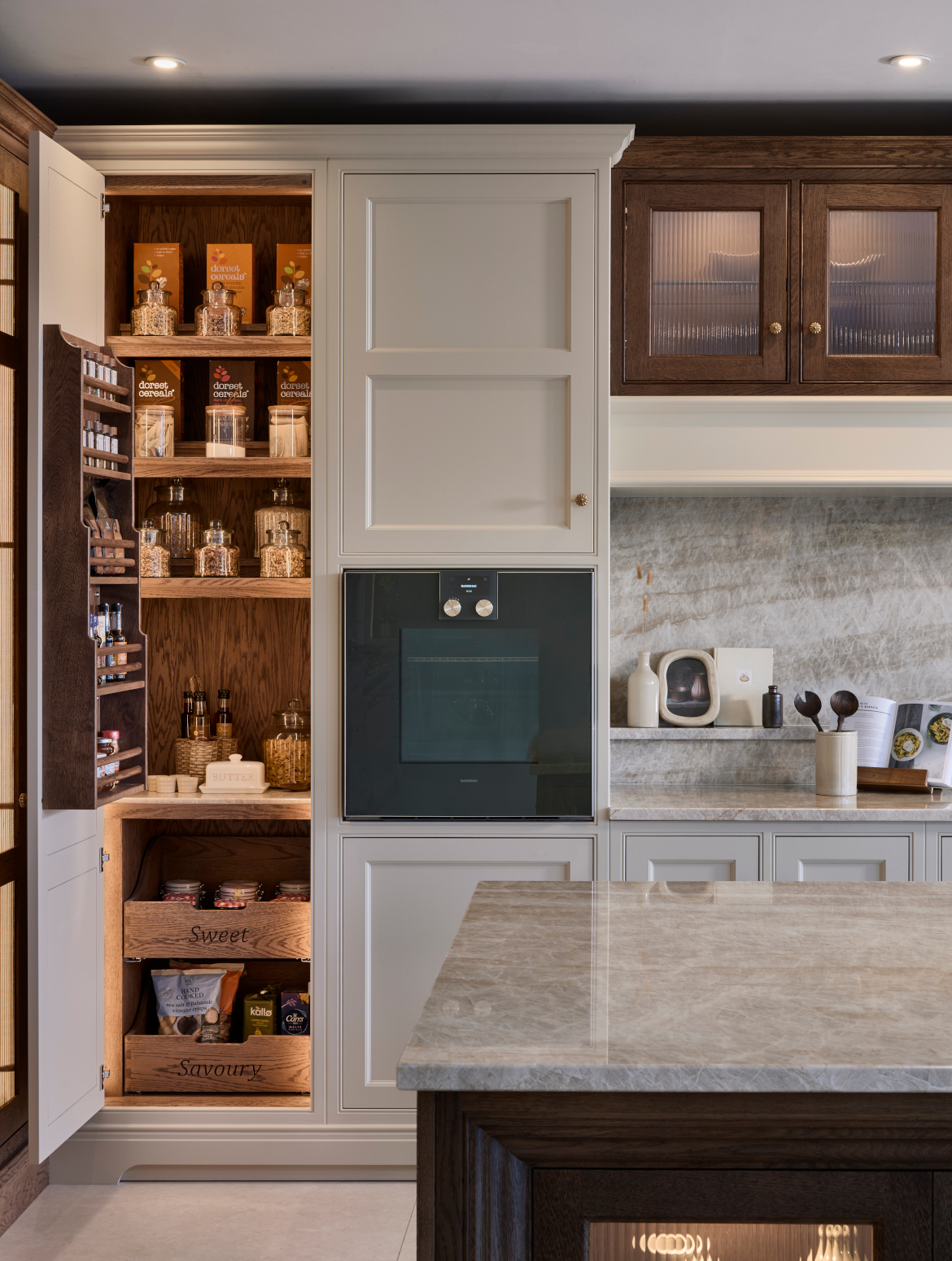 A traditional shaker kitchen in French Grey. A bespoke island stands centrally. The Taj Mahal stone stands out from the Stony Ground colour of the bespoke cabinetry beyond. Beyond the island, stands bespoke cabinetry surrounding the mantel. A Gaggenau oven stand proud in the middle of the cabinet. Whereas to the left of it, the open door reveals a pantry. The well-stocked shelves feature cereals, pasta, spices, and below there are two labelled drawers: sweet and savoury. 