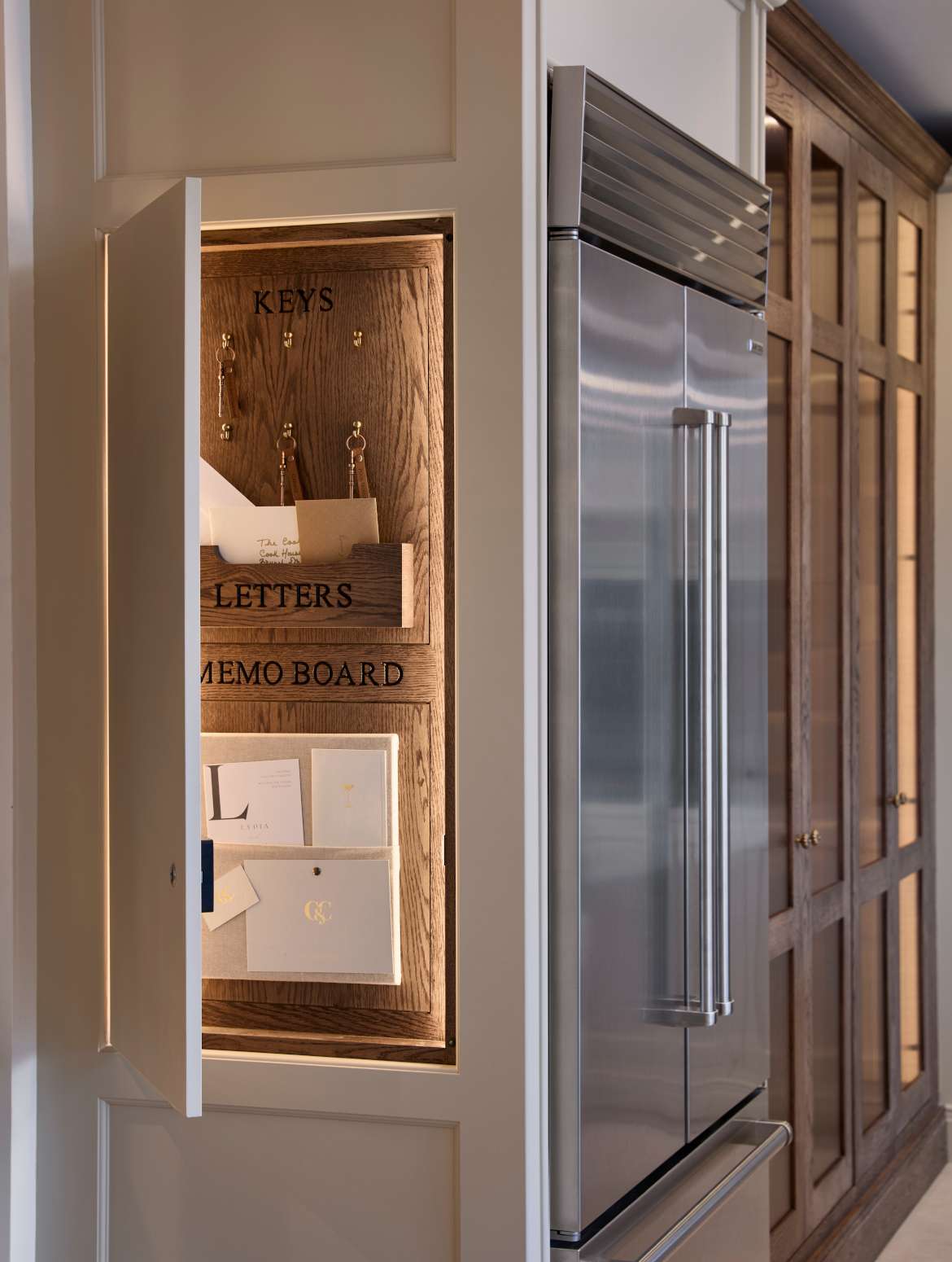A traditional shaker kitchen in French Grey. A Sub-Zero fridge freezer forms a corner with a key and letter cupboard. Their parent cabinetry surrounds them and contrasts well with the wooden accents of the open cupboard. There are key hooks above a letter shelf and memo board. 
