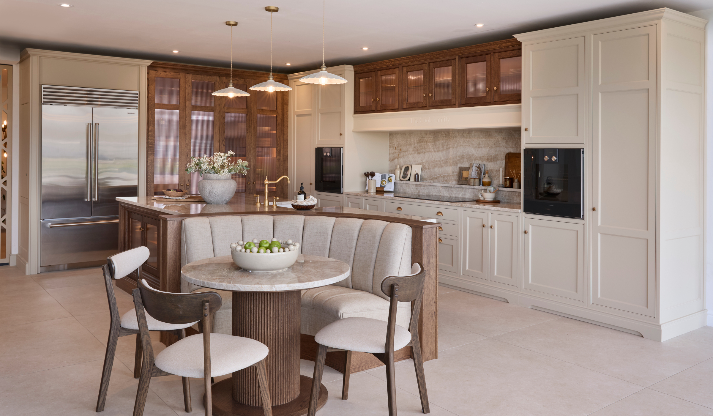 The Cooks Family Traditional Shaker Kitchen Project A traditional shaker kitchen in French Grey. A bespoke island with a seating area embedded within its curved edge stands centrally with three upholstered chairs around it. The Taj Mahal stone stands out from the Stony Ground colour of the bespoke cabinetry beyond and adjacent. To the right side of the island stands bespoke cabinetry surrounding the mantel. Two Gaggenau ovens stand proud on each side. Beyond the island, the floor-to-ceiling pantry and Sub-Zero fridge freezer stand next to each other. Armac Martin handles around the kitchen provide a further contrast to the Taj Mahal marble.