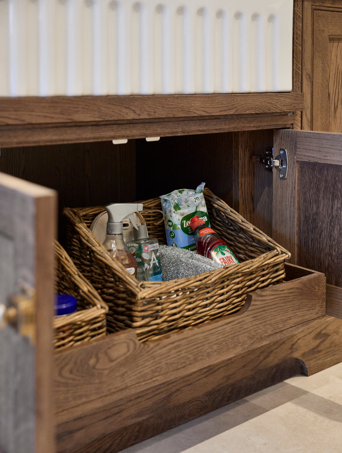A traditional shaker kitchen in French Grey. A bespoke under-sink open cupboard reveals two wicker baskets. The completely visible basket has a selection of cleaning products. 