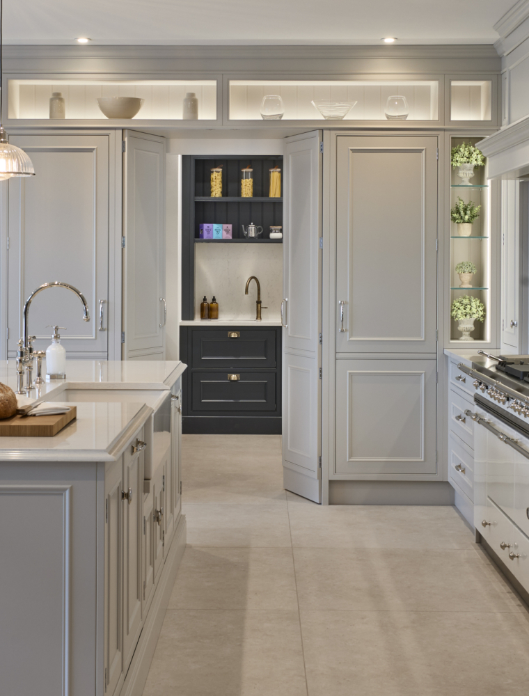 A traditional shaker kitchen in French Grey. The mantel and bespoke cabinetry surrounding a white Lacanche range cooker guides the eye to the open pantry doors ahead. While the bespoke central island fitted with Snowy Ibiza quartz catches light from three hanging bulbs above. A chopping board with sourdough loaf sits beside the sink.    