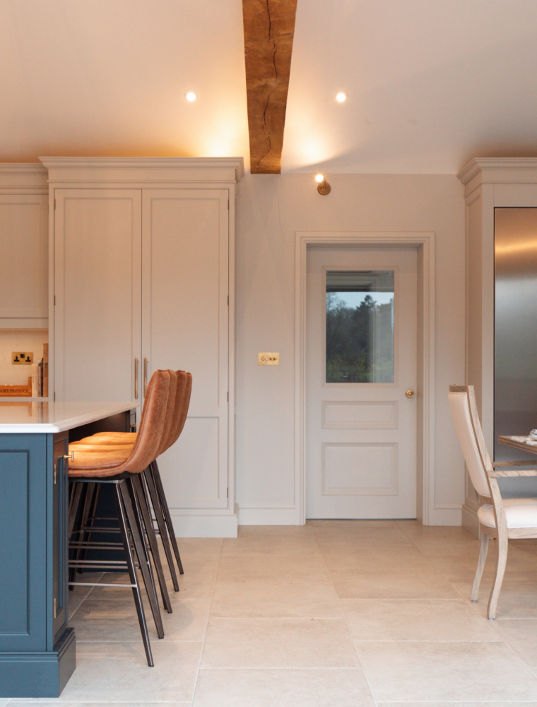 A traditional shaker kitchen in French Grey. An off-black island stands left of bespoke cabinetry set against the back wall. One side the kitchen has pantry and other bespoke cupboards and drawers. The other side houses a Liebherr monolith refrigeration bank that oversee a dining table set for a meal. Warm light above emphasises the exposed beams above.