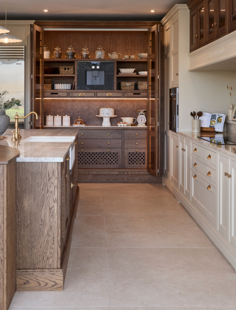 A traditional shaker kitchen in French Grey. A bespoke central island with a seating area embedded within its curved right edge. The Taj Mahal stone stands out from the Stony Ground colour of the bespoke cabinetry beyond. Two floor-to-ceiling cabinets house Gaggenau ovens while the bespoke mantel overlooks a frameless induction hob. To its left, a pantry a Sub-Zero fridge overlook the central island fitted with Bowland Shaw 600 Sink. 