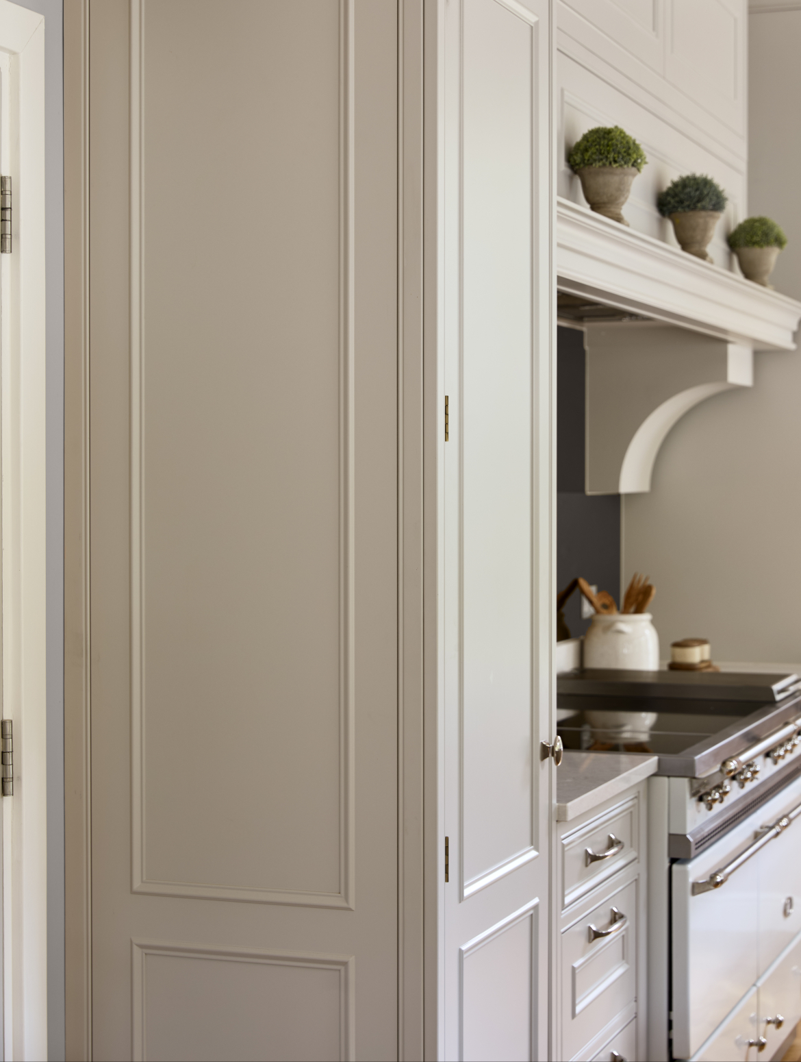 A traditional cream shaker kitchen with bespoke cabinetry showcasing the flush edges of a ceiling-high cabinet that stands as part of the cabinetry over a white Lacanche range cooker. It also illustrates the joints and hinges of the bespoke piece. 