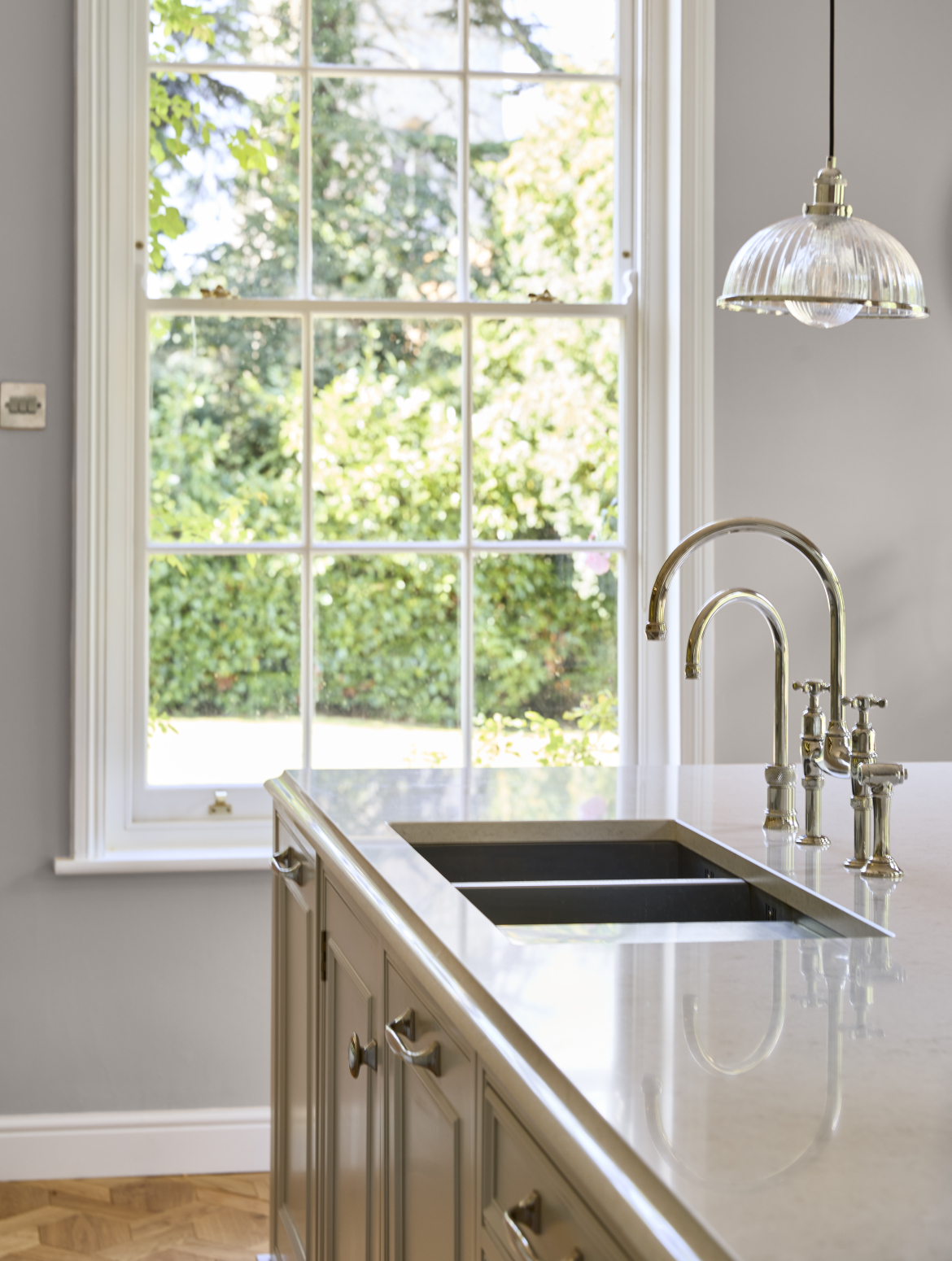 A traditional cream shaker kitchen a central island and bespoke cabinetry. The island has a Quooker and a Perrin and Rowe tap with cupboards and drawers below. This corner of the island is overlooked by a large Georgeian-style window showing a variety of trees and bushes outside. 