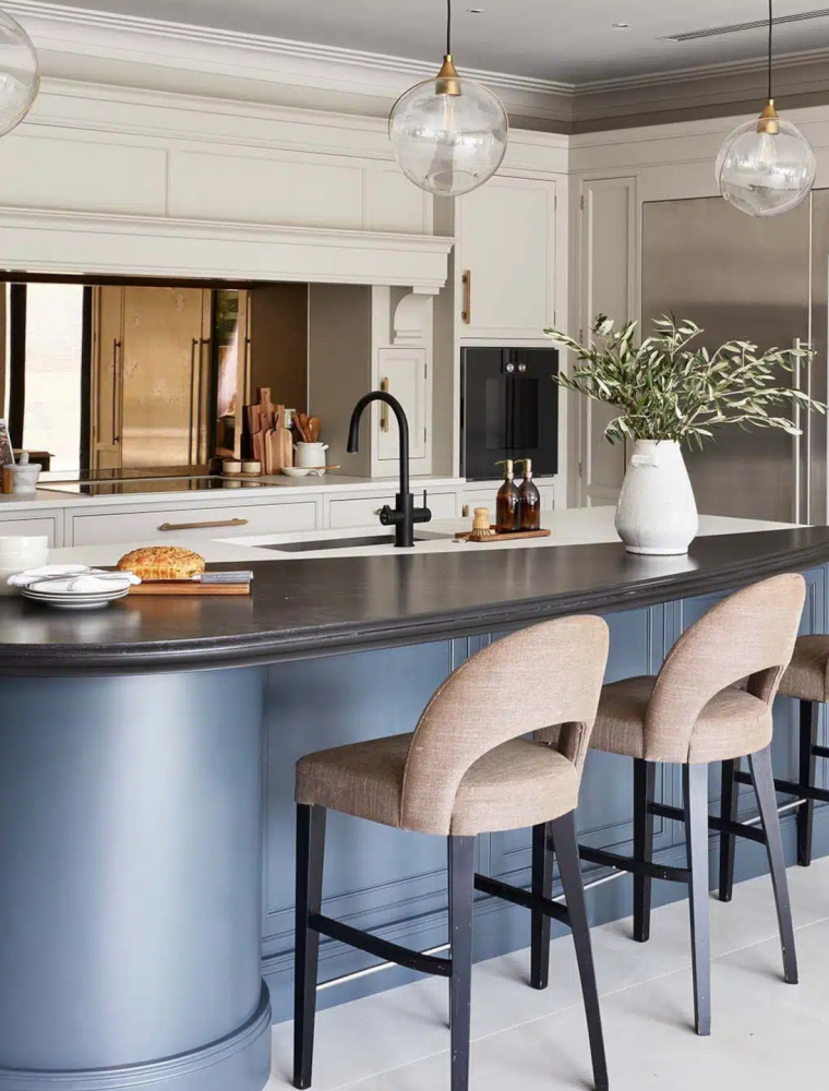A traditional shaker kitchen project in off-black. A central island with the bespoke cabinetry and a streamlined wood ledge overseeing three upholstered highchairs contrast the white worktop of the remaining island beyond. To its left, bespoke off-black cabinetry oversees the cabinetry opposite that houses Liebherr monolith fridge and freezer. To their right, two Gaggenau ovens are built-in to the middle French Grey cabinetry. 