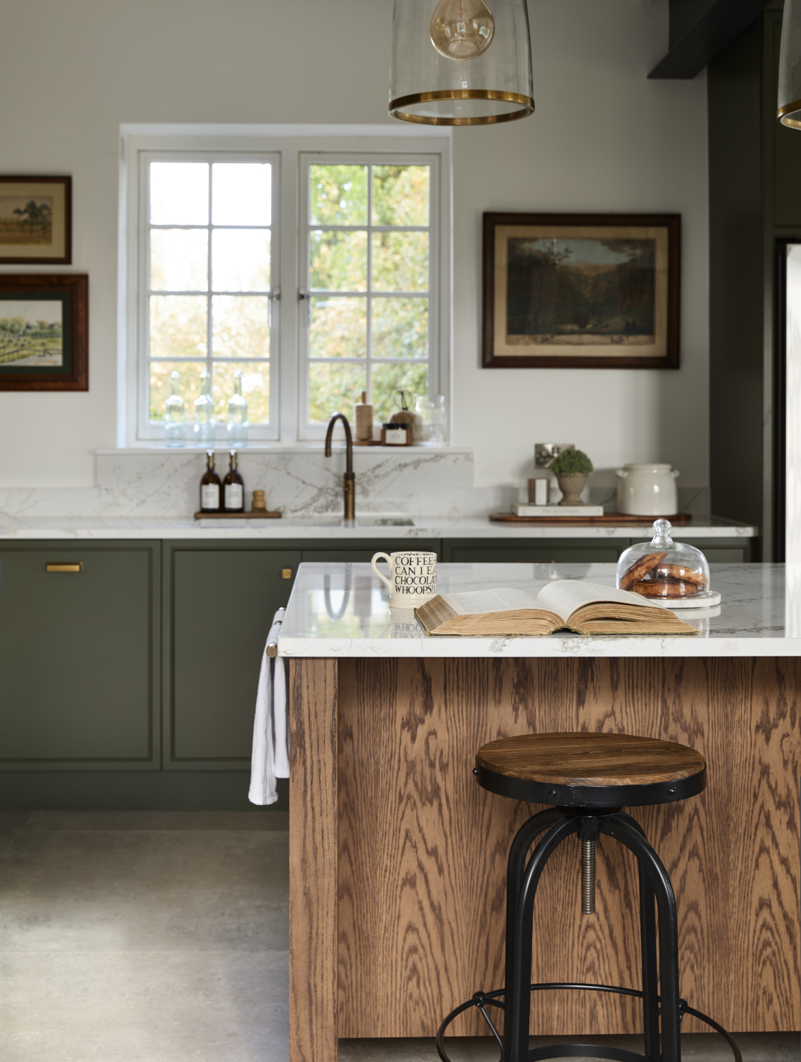 A traditional shaker kitchen in Invisible Green. A view of the raised central island’s Verde Peak marble edge. The lip oversees a black-legged stool with wooden seat. On the marble top, an open book lies before a mug and a covered plate of three pain au chocolats. Though in the background, the patinated Quooker Fusion tap stands prominently on the worktop beyond. 