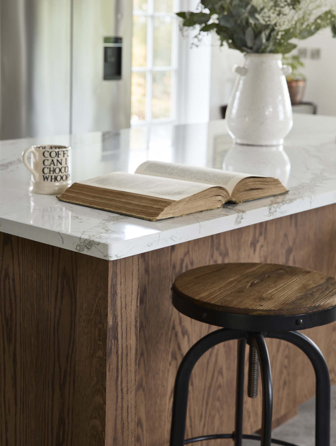 A traditional shaker kitchen in Invisible Green. A close in view of the raised central island’s Verde Peak marble edge. The lip oversees a black-legged stool with wooden seat. On the marble top, an open book lies before a mug. 